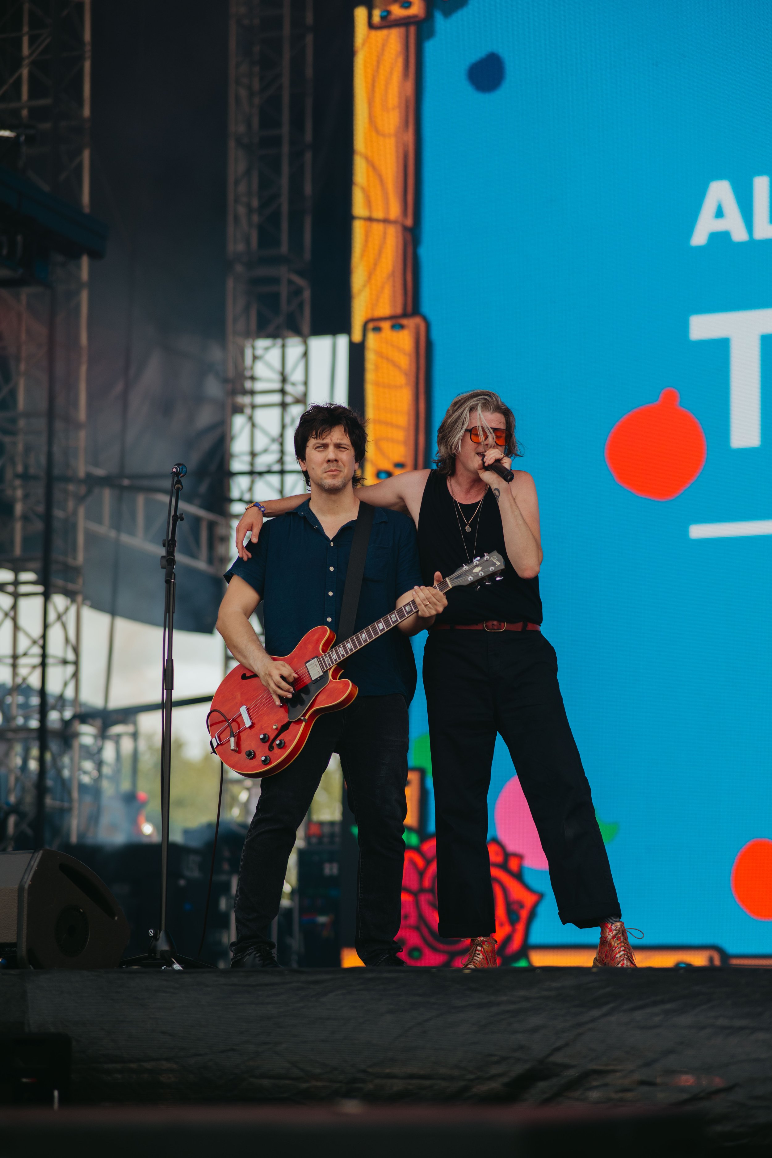 Two musicians performing on stage: a man with dark hair playing a red electric guitar and a woman with blonde hair wearing sunglasses singing into a microphone. They stand in front of a large, vibrant digital screen with colorful graphics.