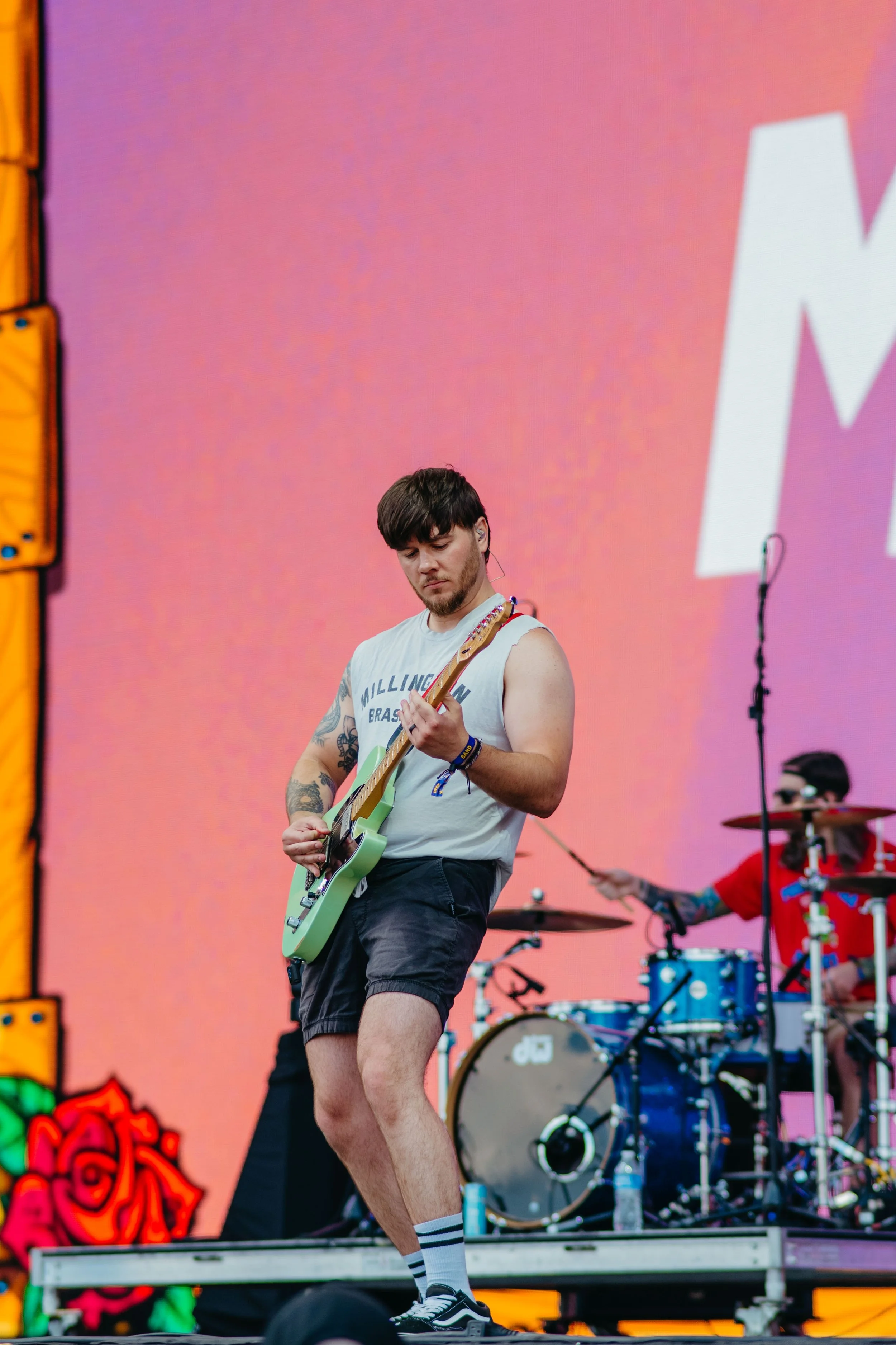 A young male guitarist on stage playing an electric guitar, wearing a sleeveless shirt, shorts, and striped socks, during a band performance with a drummer in the background against a colorful digital screen backdrop.