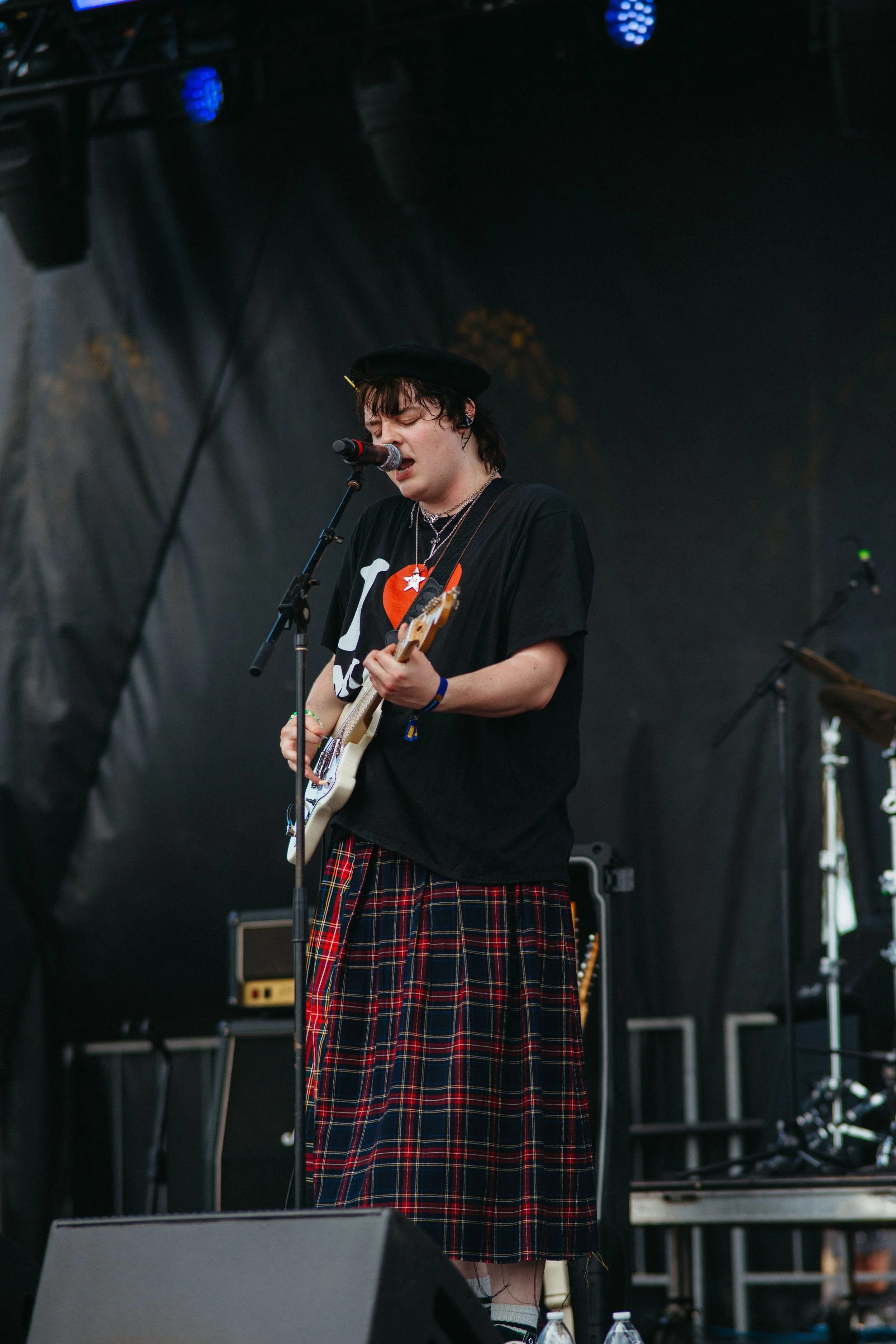 A young person with dark, messy hair singing into a microphone while playing an electric guitar on stage. They are wearing a black beret, a black T-shirt, multicolored jewelry, and a traditional tartan skirt. The stage has various equipment and cable
