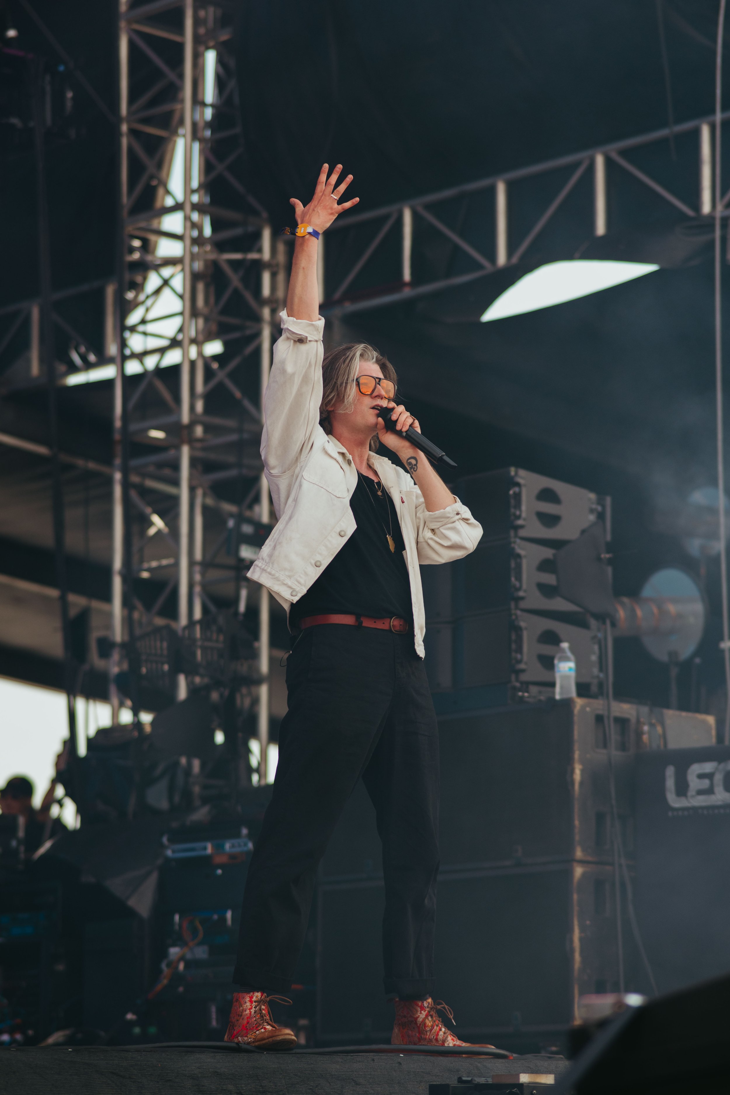 A person with shoulder-length hair, wearing sunglasses, a white jacket, black pants, and red patterned boots, is standing on stage, holding a microphone, with their right arm raised.