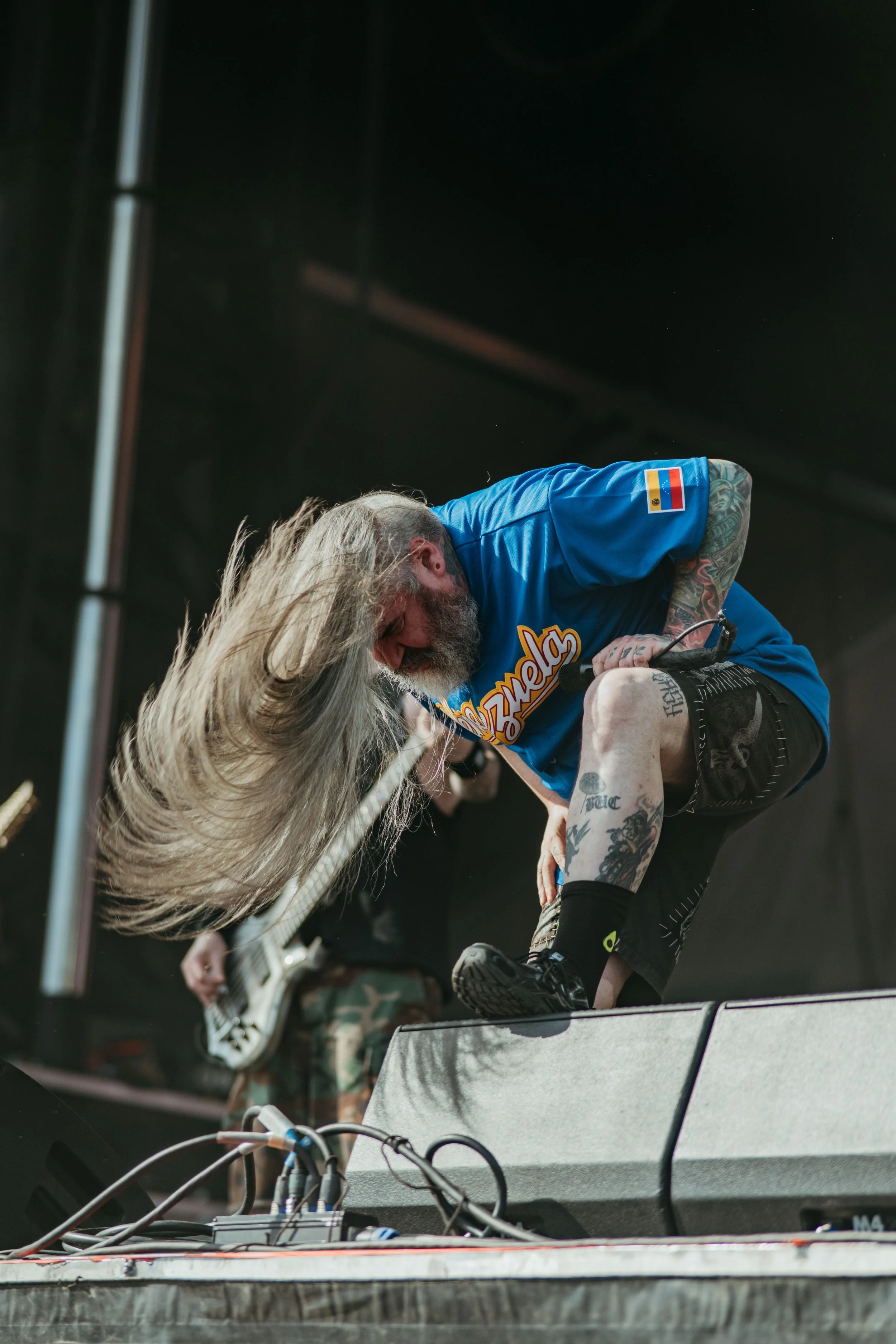 Muscular man with tattoos wearing a blue jersey and black shorts is on stage, performing with a musician on guitar, in a concert setting with equipment in the foreground.