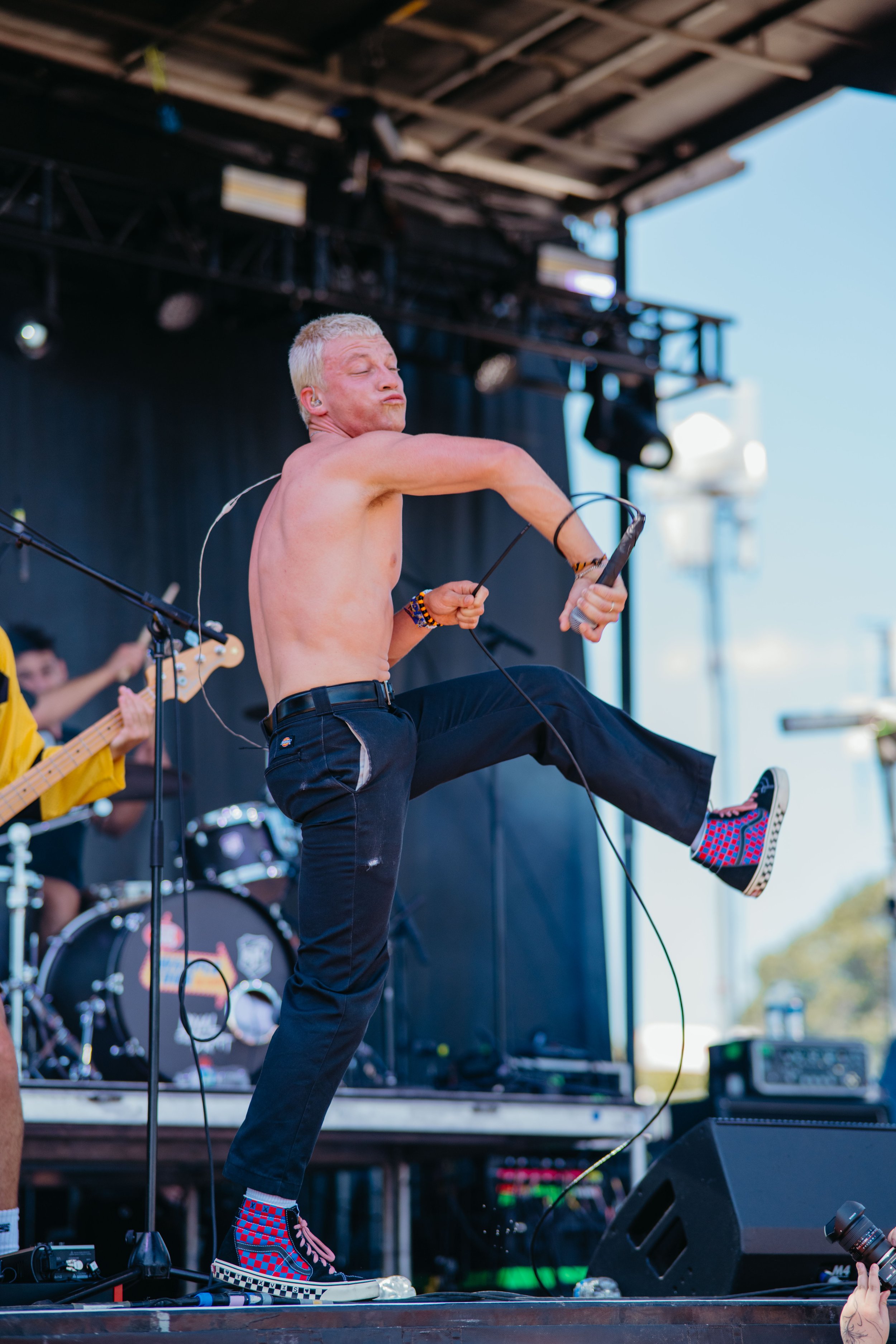 A shirtless man with blonde hair dancing on stage during a live music event, with a guitar, drums, and stage equipment visible in the background.