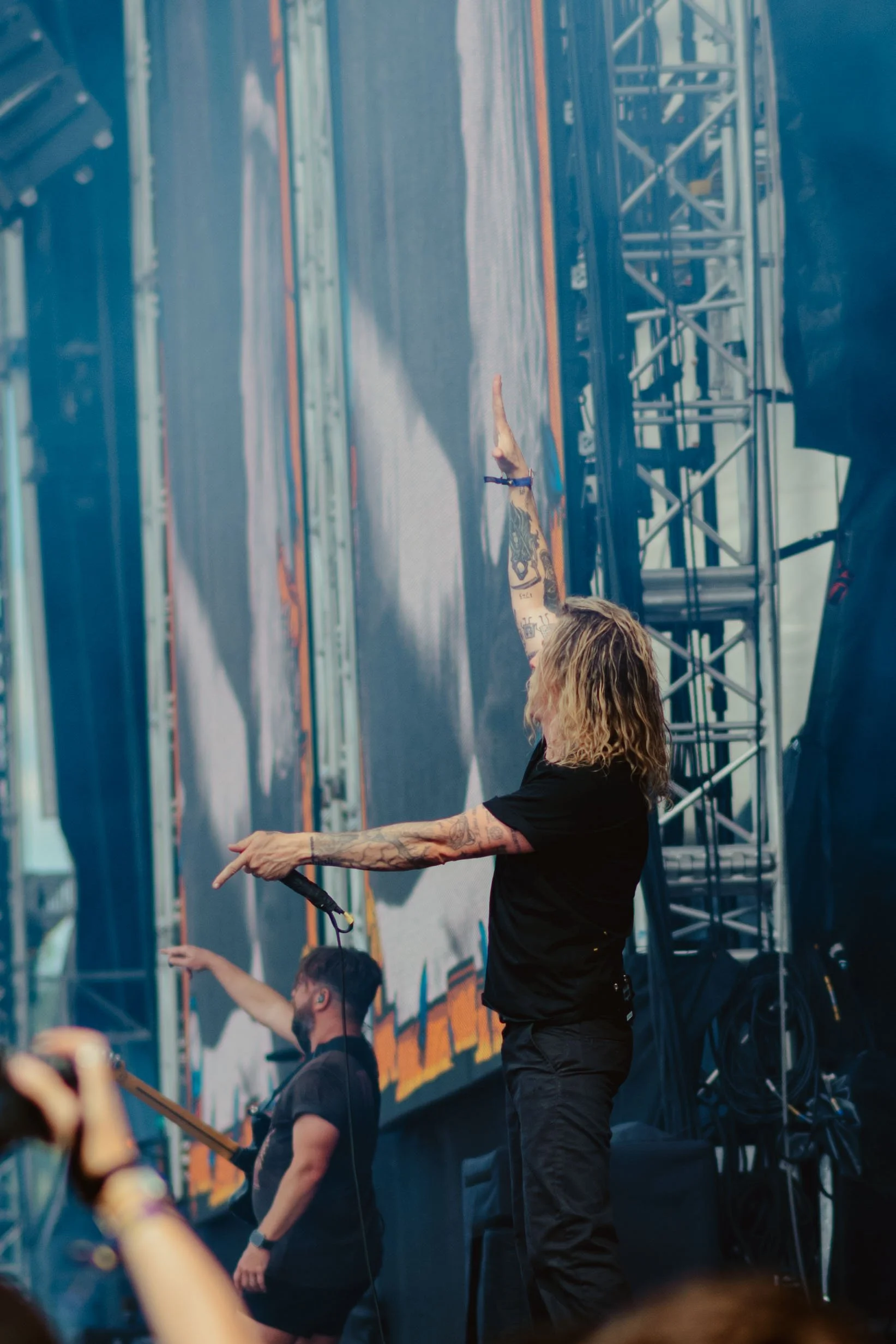 Two musicians perform on stage during a concert. One has tattoos and is raising an arm, while the other is wearing glasses and using drumsticks. There is a large screen in the background with vibrant visuals.He is in the band Underoath.