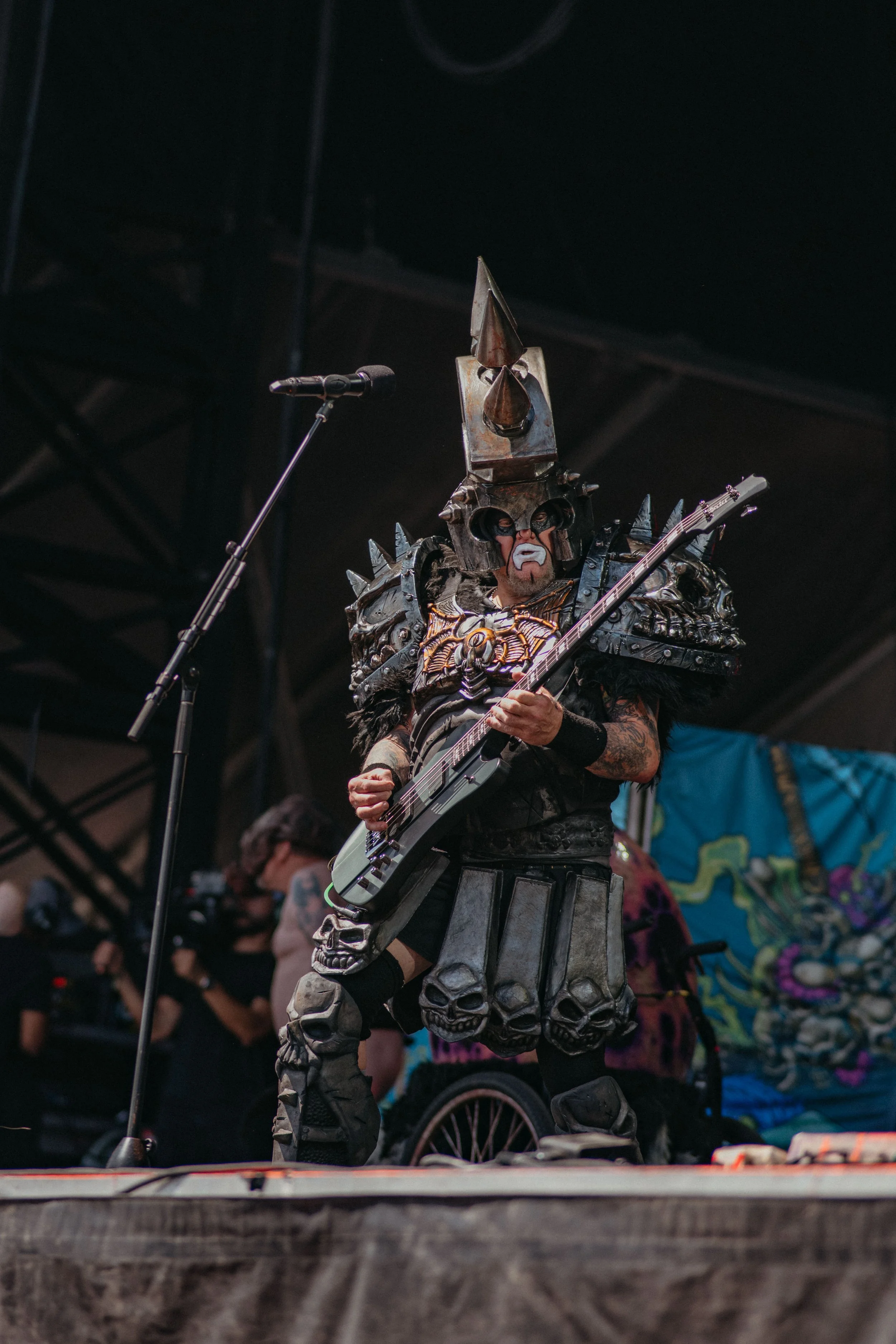 A musician dressed in elaborate, medieval-inspired armor with spikes and skull motifs, playing an electric guitar on stage with a microphone nearby.