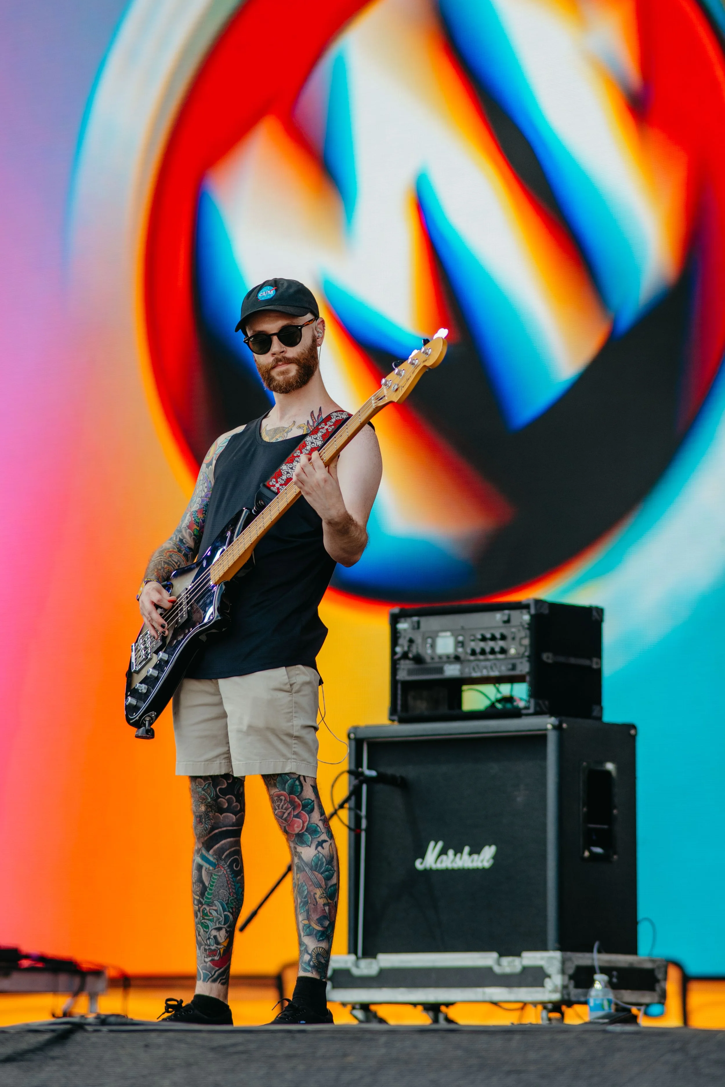 A tattooed man wearing sunglasses, a black tank top, beige shorts, holding a guitar on stage with a large colorful abstract background, and a Marshall amplifier in front of him.