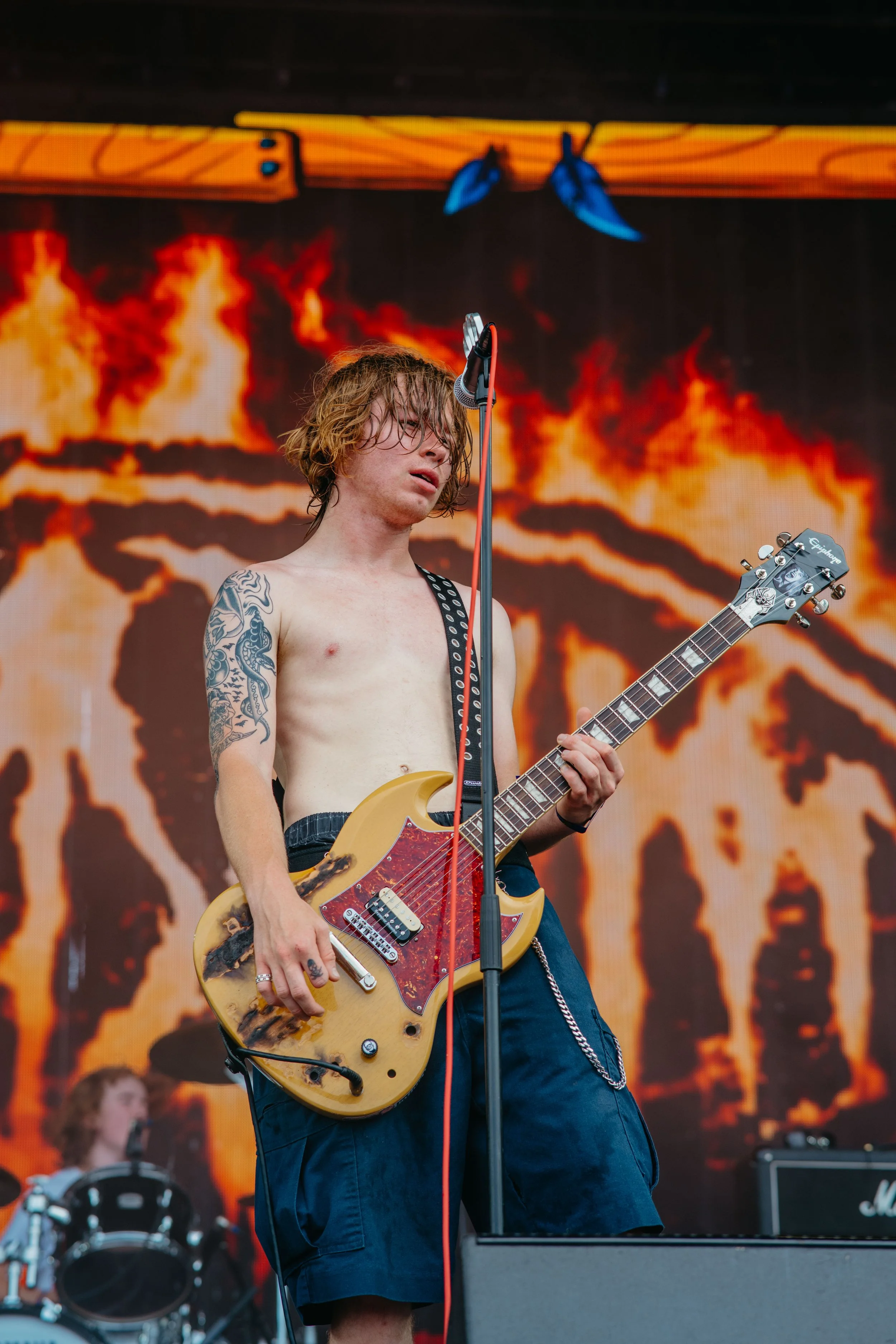 A shirtless young man with tattoos playing an electric guitar on stage at a music concert, with a fiery background on a large screen.