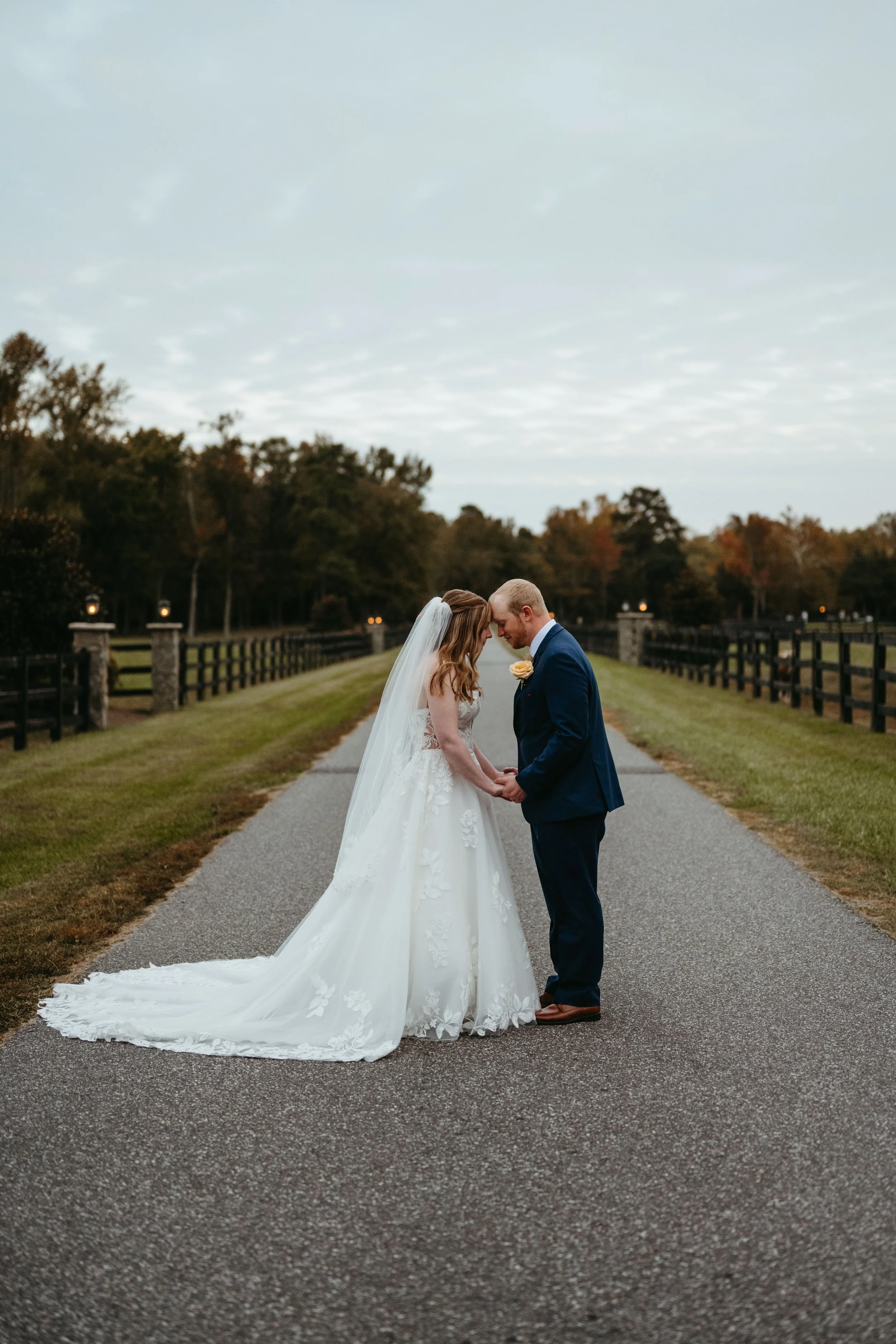 A bride and groom holding hands and touching foreheads in the middle of a paved road, with trees and a fence on either side, during their wedding ceremony.