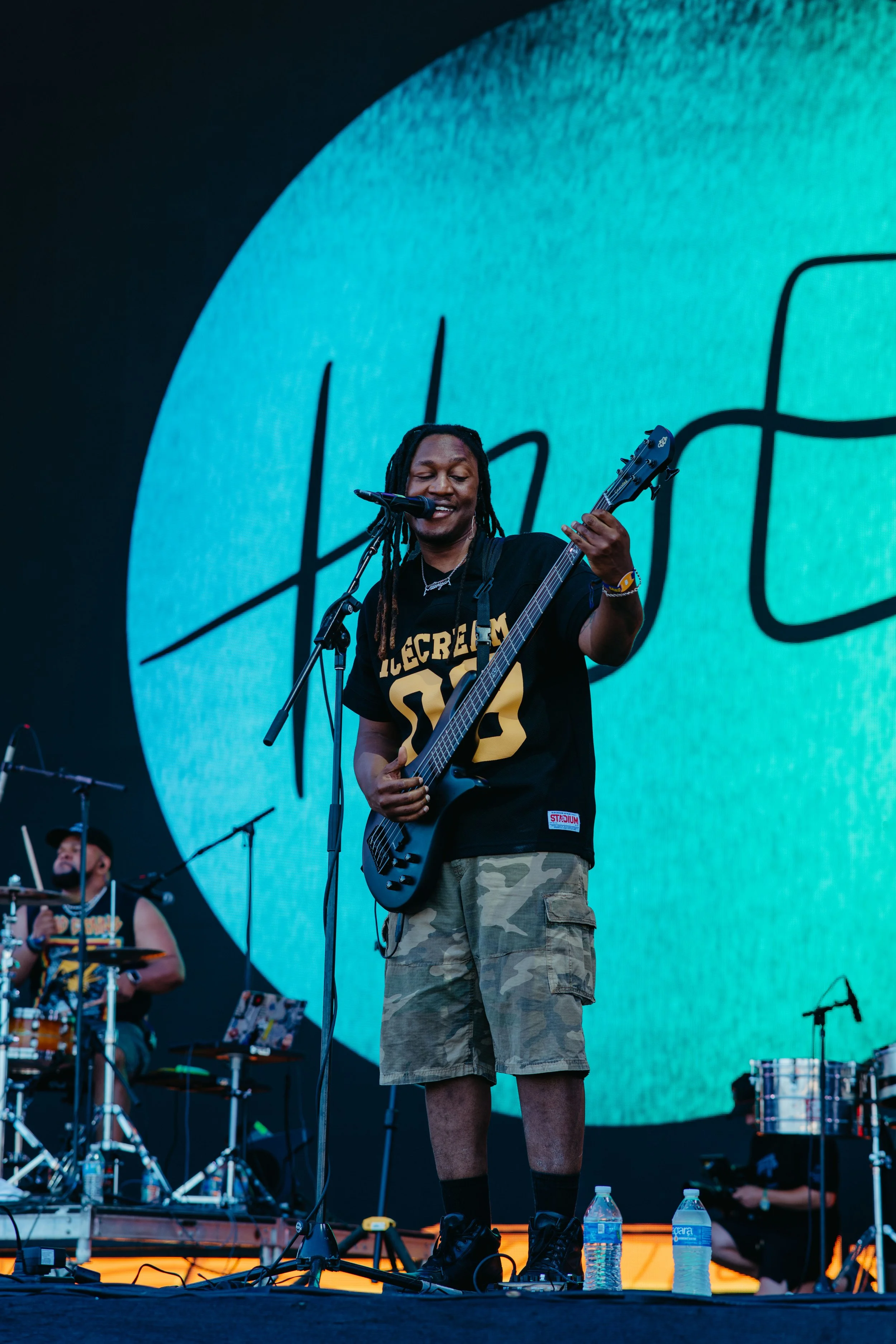 A musician with dreadlocks playing an electric guitar on stage, wearing a black T-shirt, camouflage shorts, and black shoes, with water bottles nearby. Behind him is a large digital screen with a blue gradient and black lettering, and a drummer is vi
