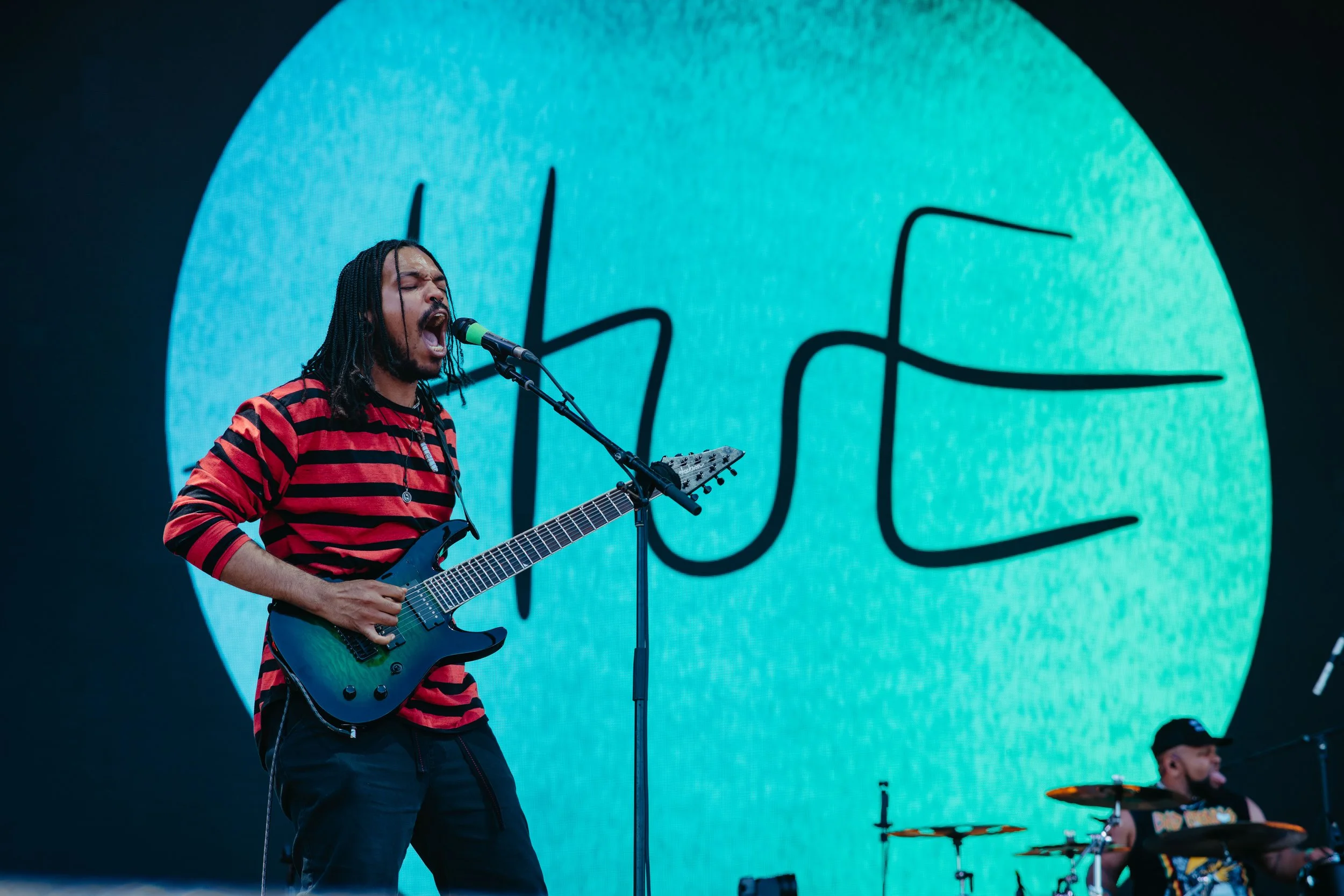 Male musician with long hair and striped shirt plays electric guitar and sings into a microphone on stage, with large screen displaying stylized smiley face in the background.
