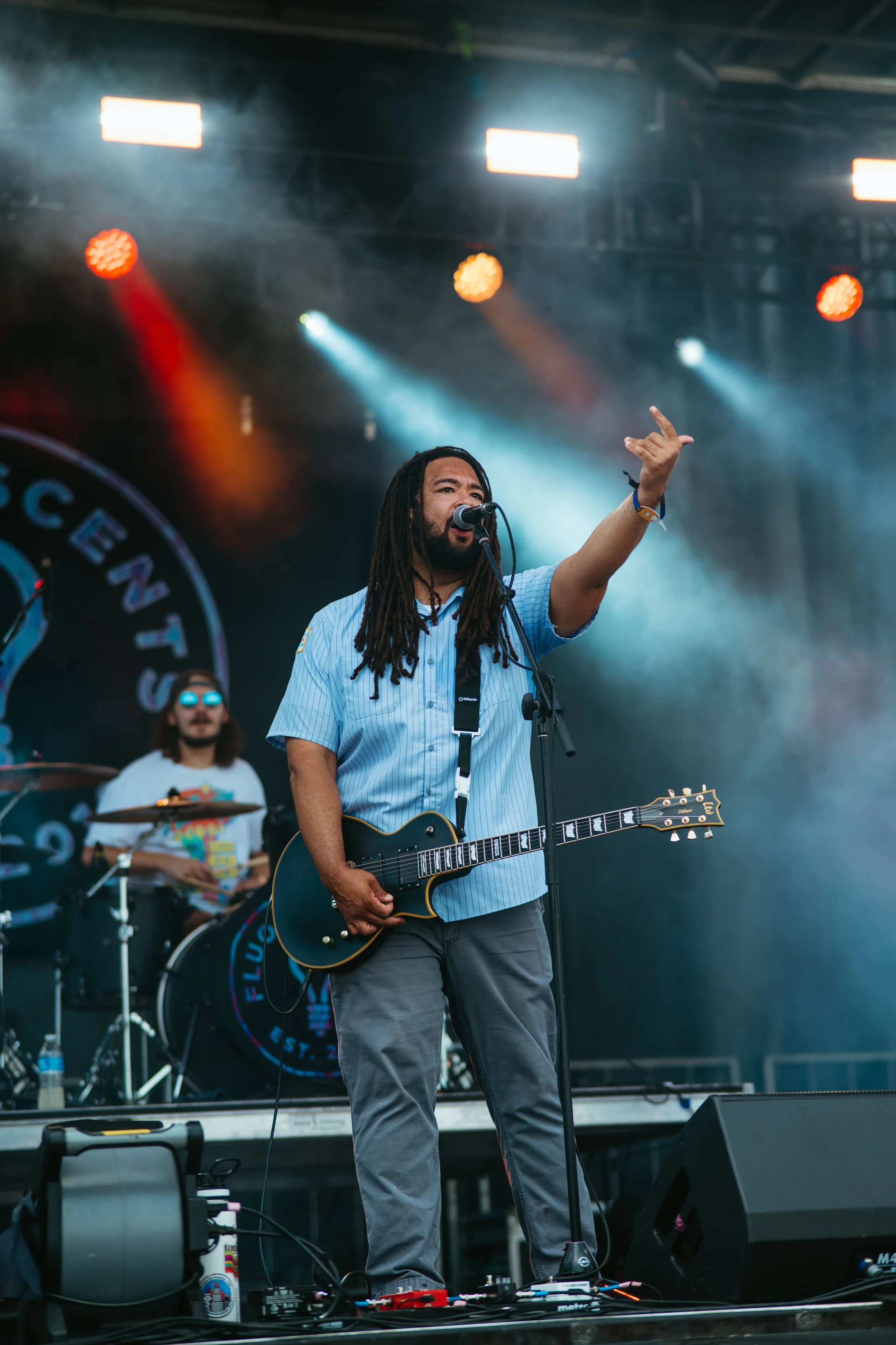 A man with dreadlocks, wearing a light blue shirt, performs on stage with an electric guitar, raising his right hand, with a drummer in the background and stage lights overhead.