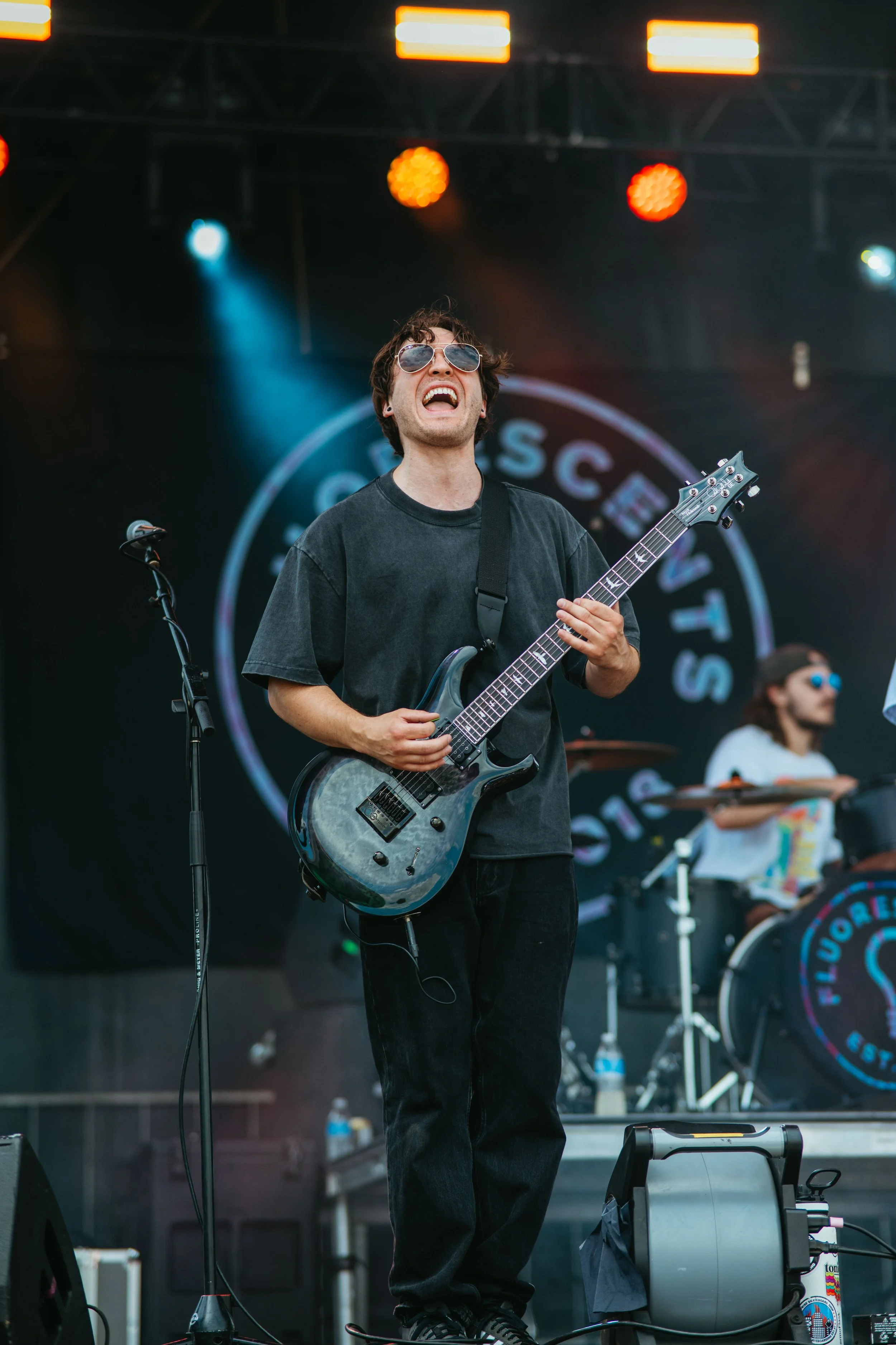 A man with sunglasses playing electric guitar during a live performance at an outdoor concert, with stage lights and a drum set in the background. He is the guitarist for the band Flourescents.