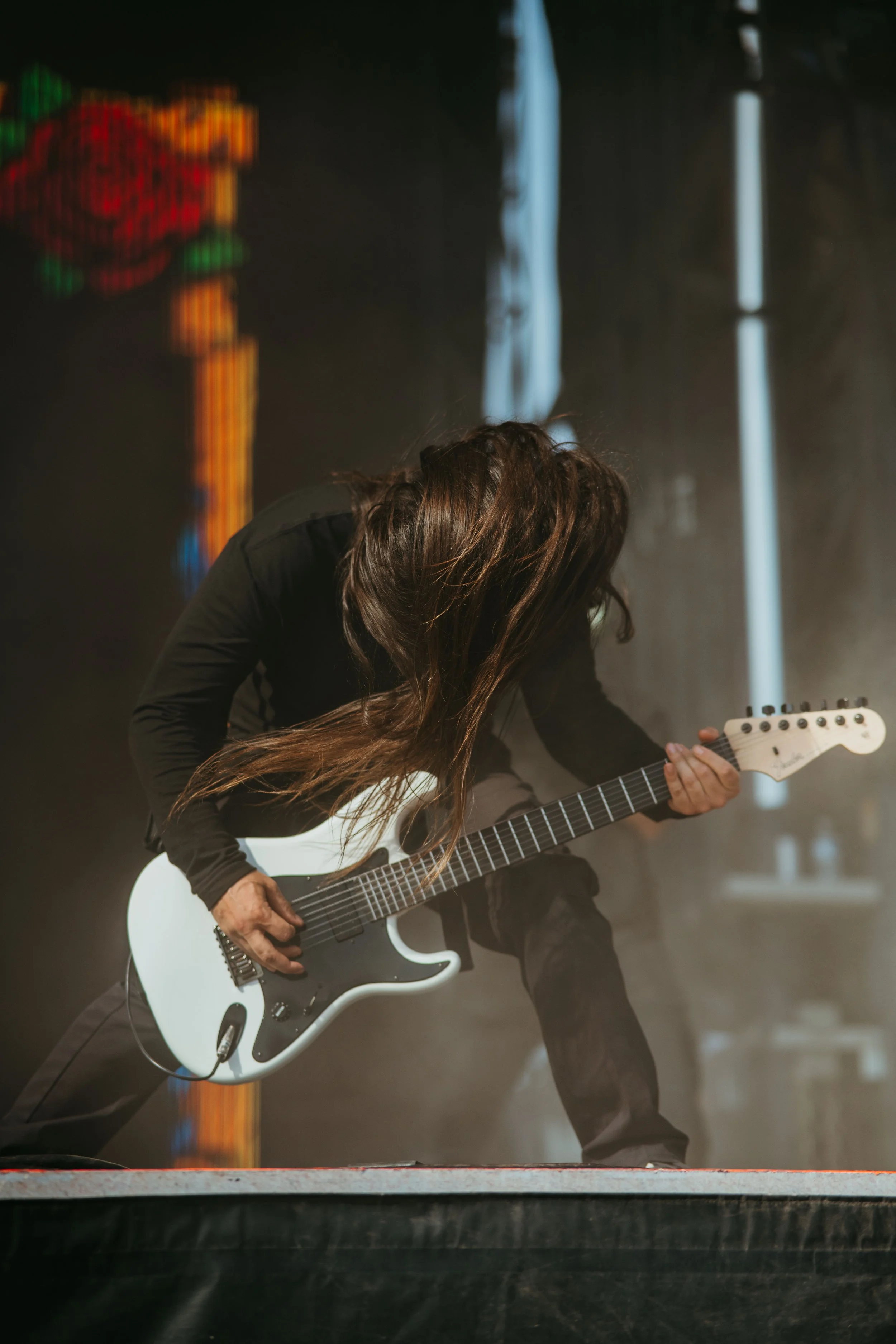 A person playing an electric guitar on stage, with long hair covering their face, standing with legs apart. The background features blurred colorful lights and vertical white light beams.