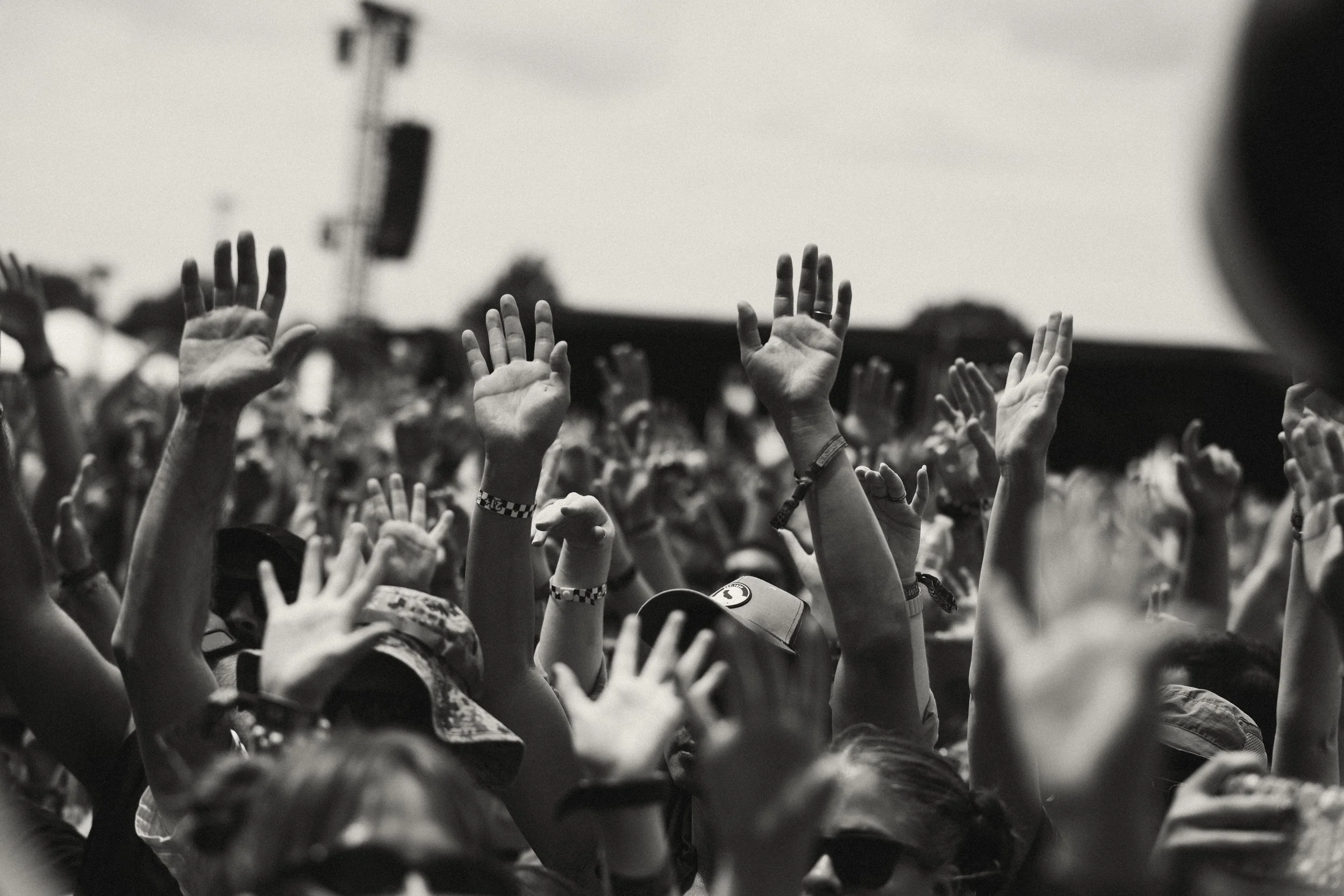 Black and white photo of raised hands at a concert or outdoor event, with a stage and equipment visible in the background.
