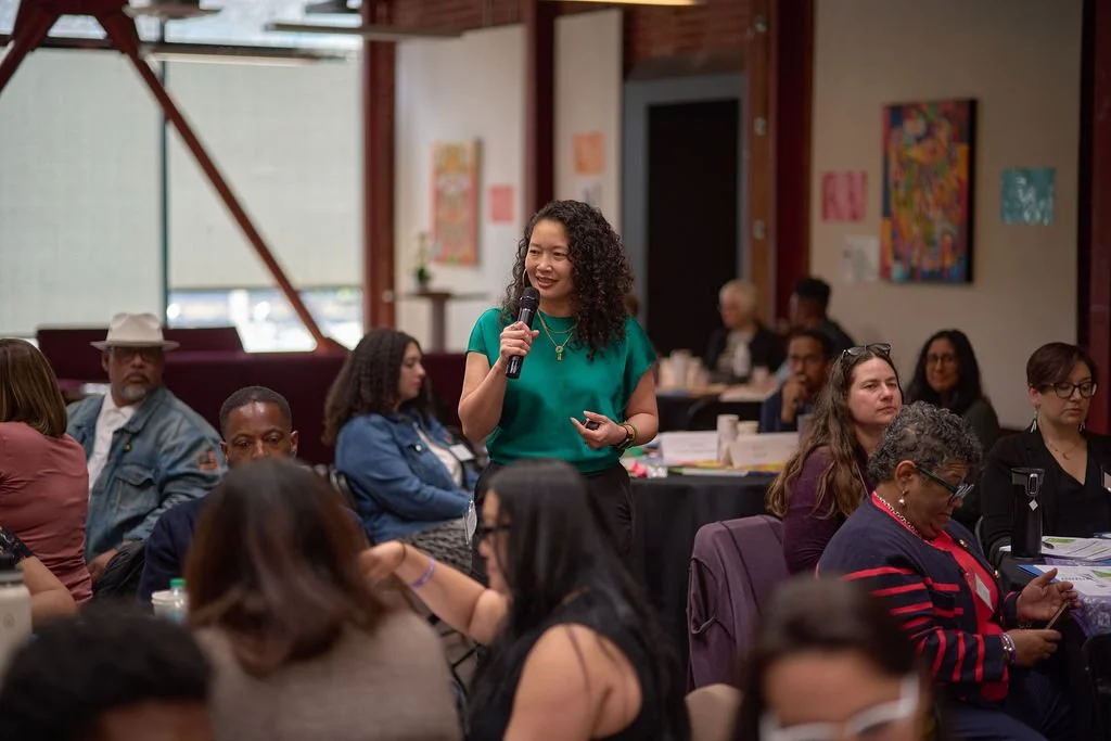 An NEP facilitator in a green shirt looks out at a room full of education leaderes.