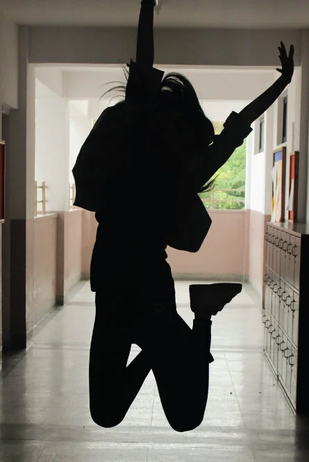 A school hallway in muted tones with a dark image in the front center of a student with long hair jumping victoriously.