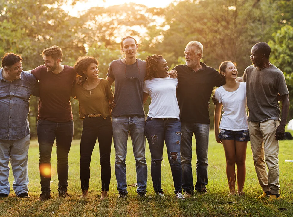 Diverse group of smiling adults with their arms around each other while the setting sun shines through the trees behind them.