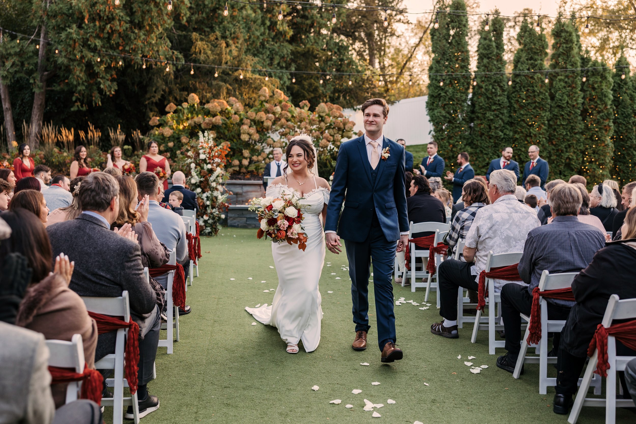 Bride and groom walking outdoors on a fall day, wearing wedding attire, with trees in the background.