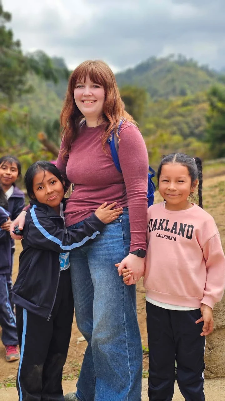 A woman with long red hair smiling while holding hands with two young girls, one girl wearing a pink sweatshirt that says Oakland California and another girl hugging the woman, in an outdoor mountainous setting.