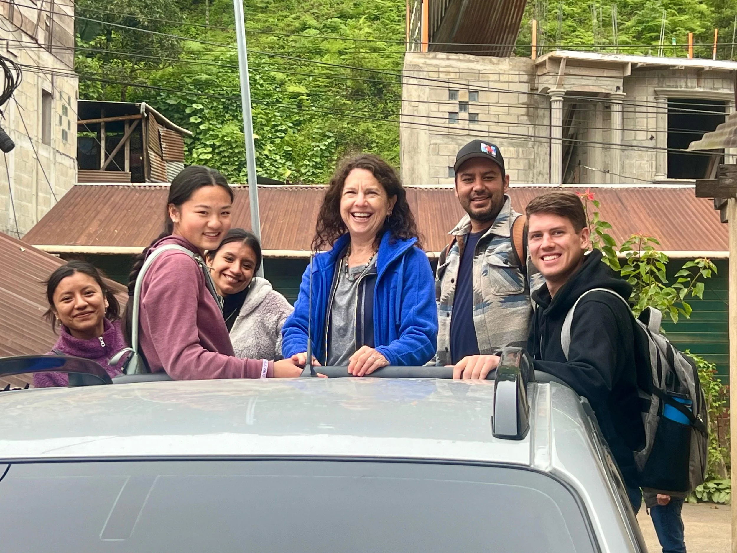 A group of six people standing behind a car, smiling outdoors with green trees and an unfinished building in the background.