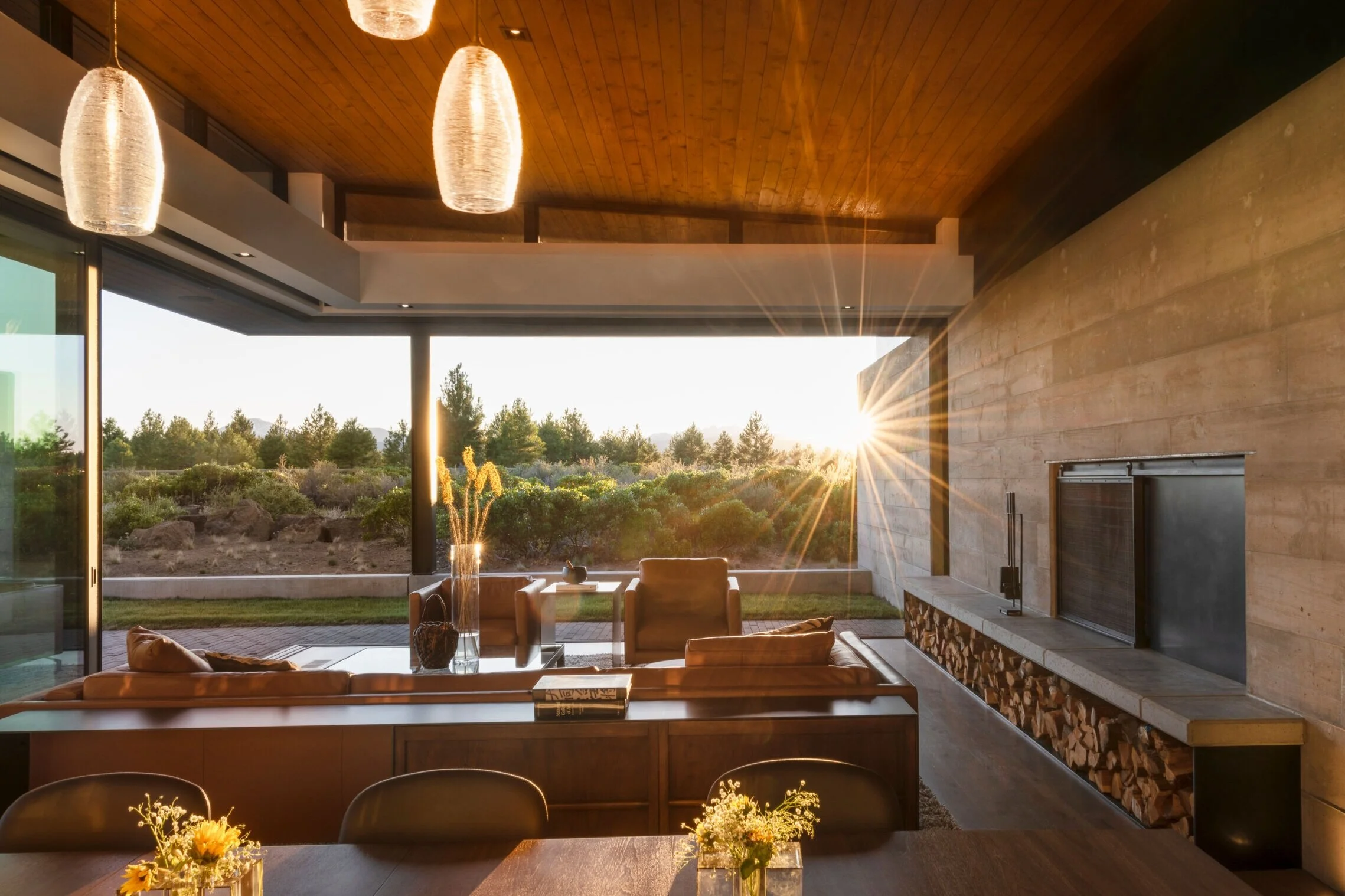 Bachelor View great room at sunset with board-formed concrete fireplace wall, firewood storage, wood plank ceiling, pendant lights, and floor-to-ceiling glass opening to high desert views in Bend Oregon