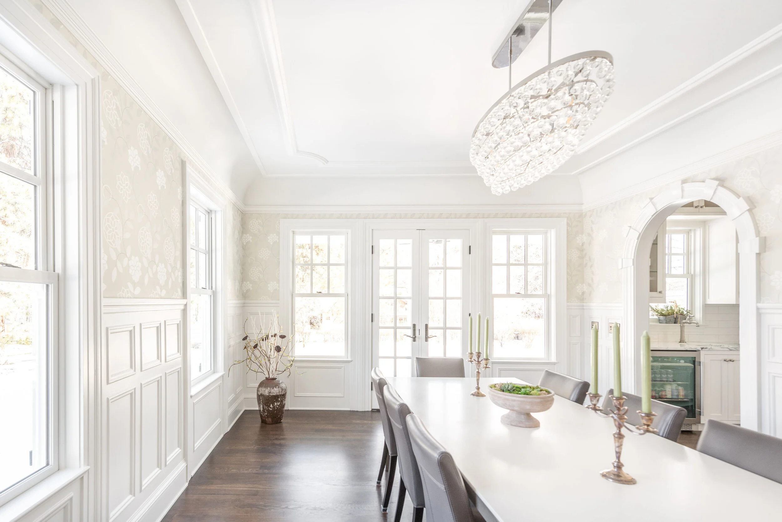 Bright dining room with white walls, large windows, a white table with silver chairs, a crystal chandelier, and decorative candles.