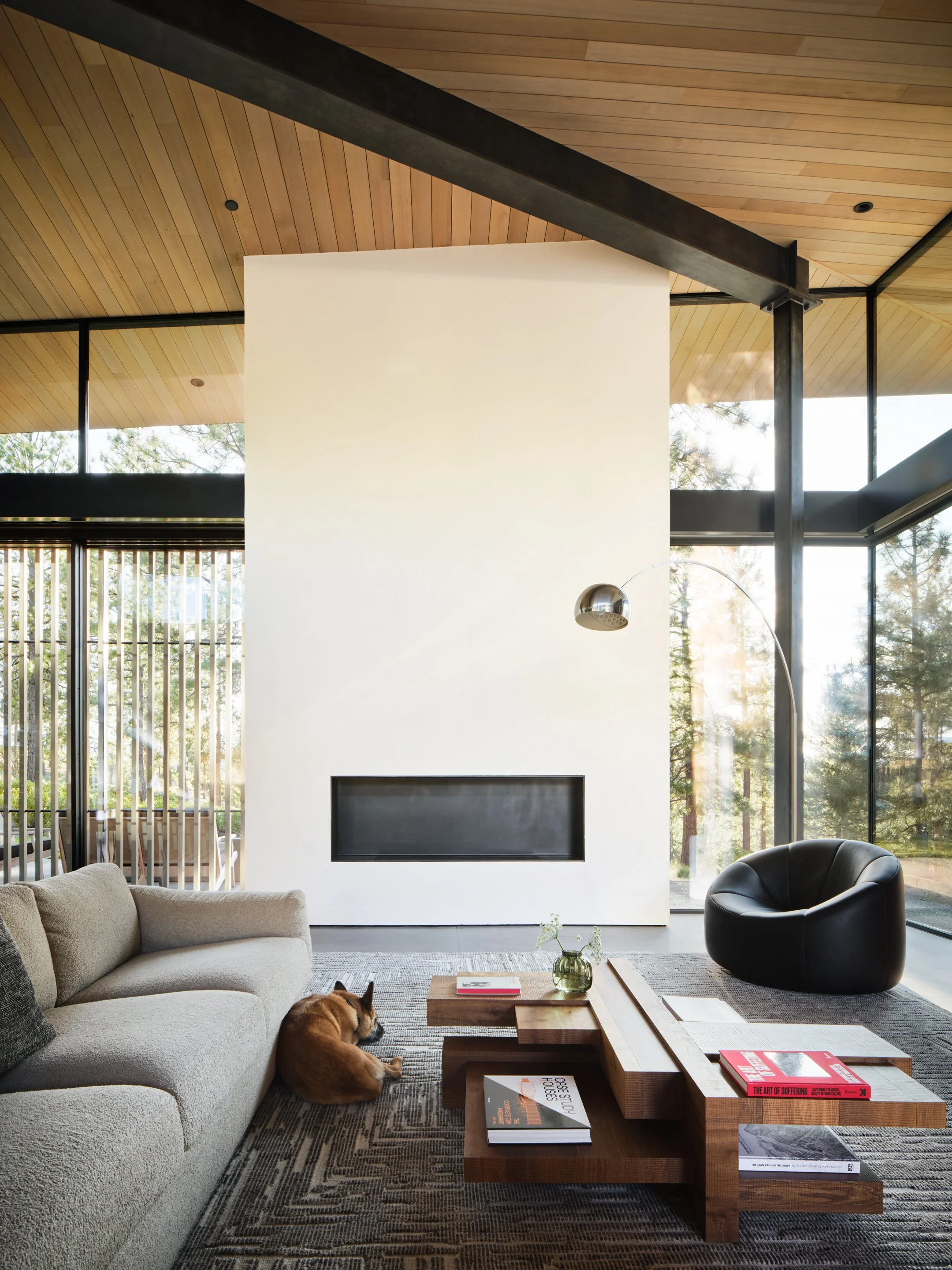 Modern living room with a beige sofa, a black leather armchair, a wooden coffee table with books, a glass vase with flowers, a floor lamp, and large windows showing a view of trees outside.