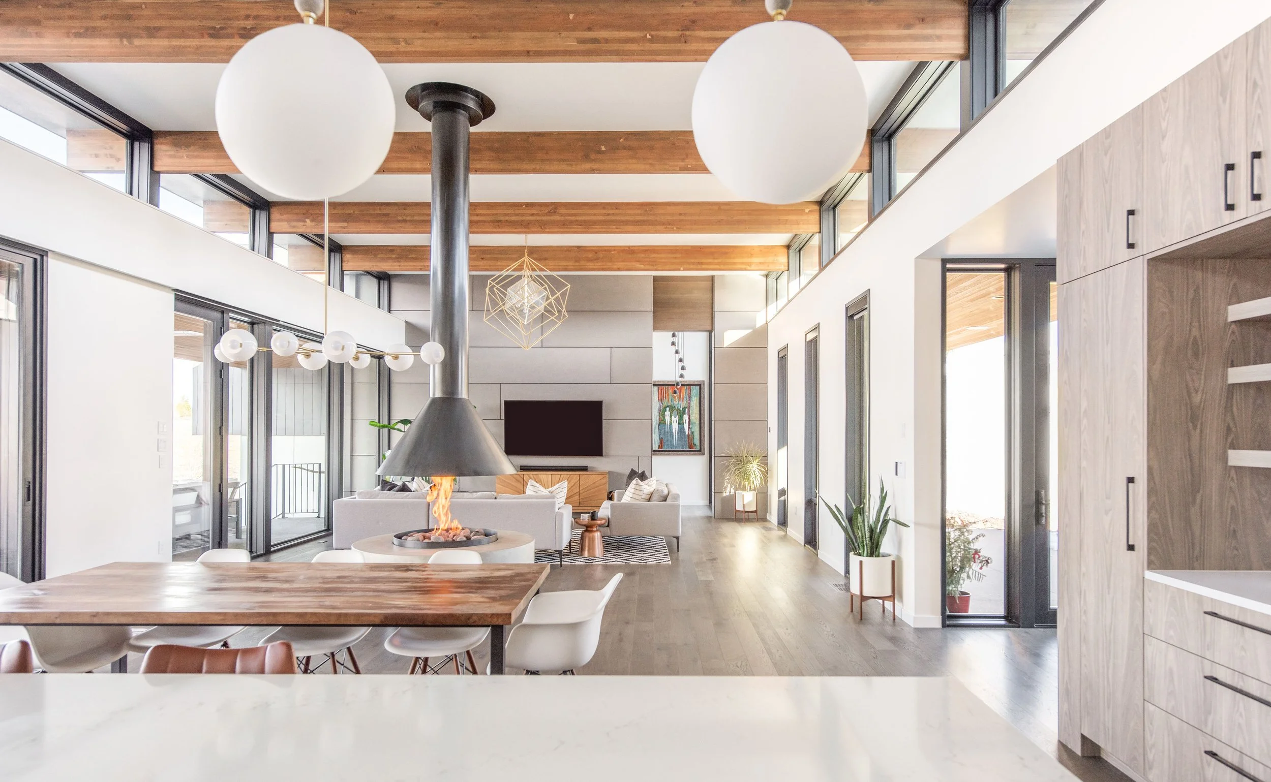 Expansive open-concept great room in a custom Bend home, featuring soaring ceilings with exposed wood beams, floor-to-ceiling windows, and a striking central suspended black fireplace.