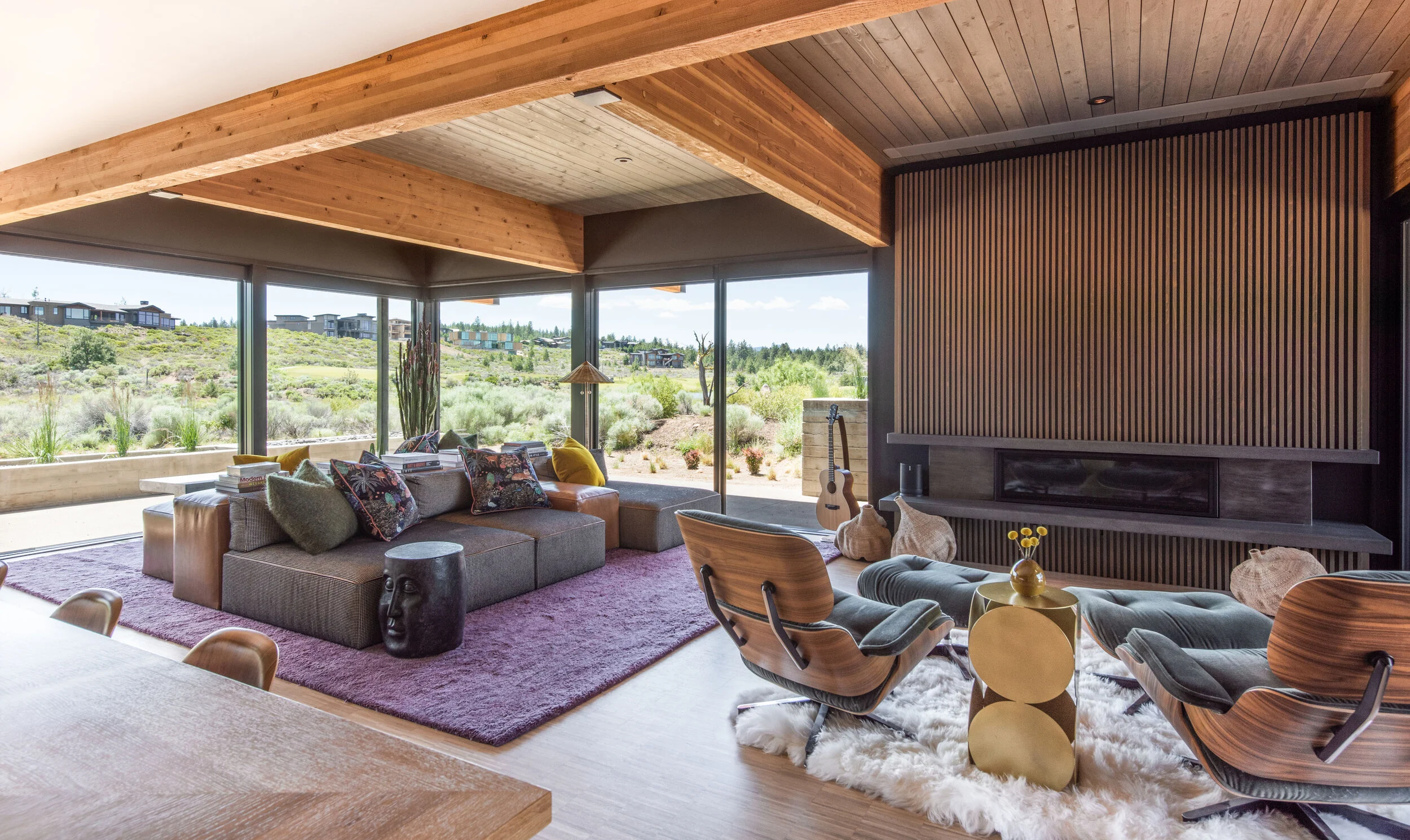Skyfall great room with exposed wood beam ceiling, vertical wood slat fireplace wall, corner windows overlooking high desert, and eclectic mid-century furnishings in Bend Oregon