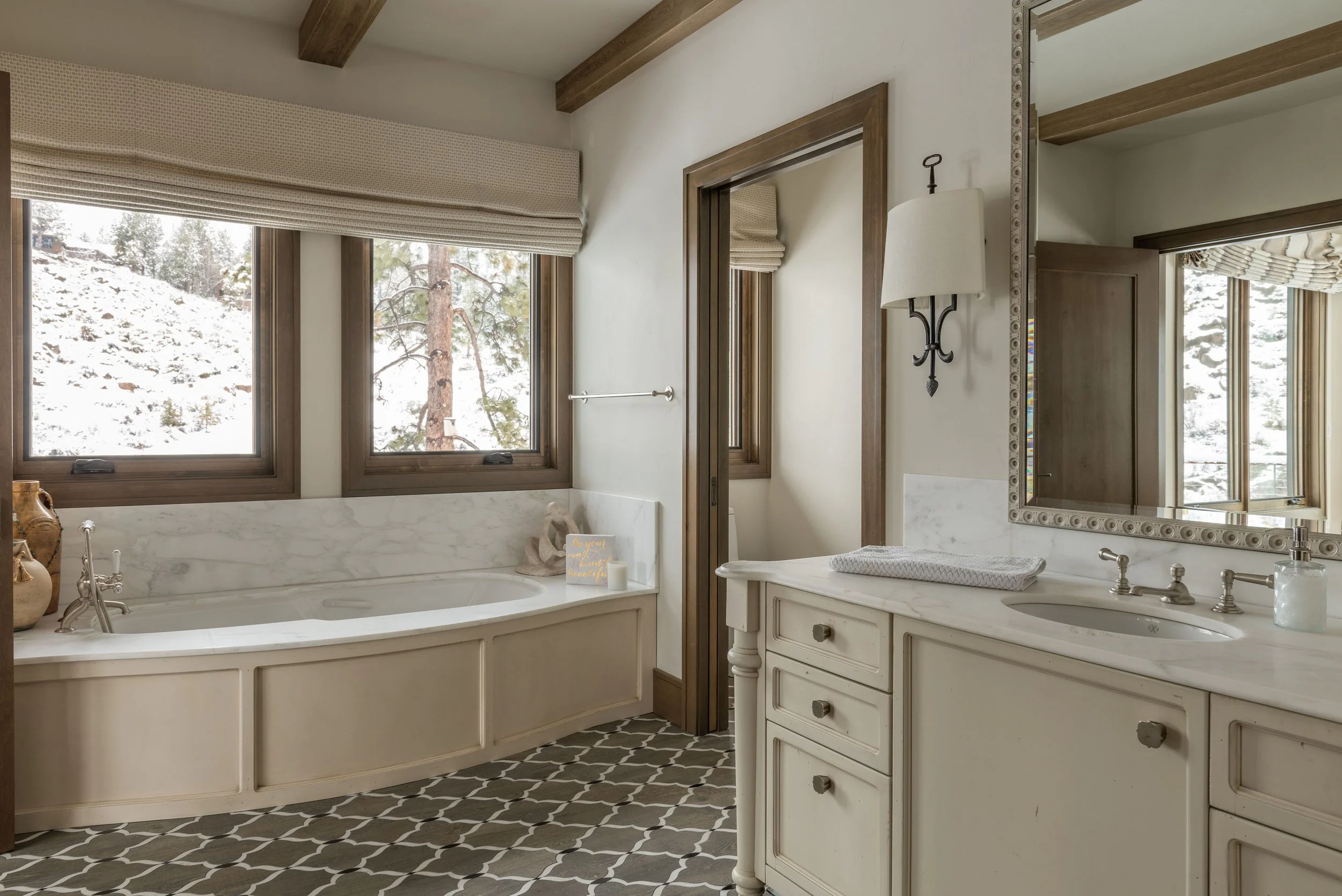 Bathroom with a large bathtub next to a window showing snowy outdoors, a white vanity with a marble countertop, and a mirror, decorated with wooden accents.