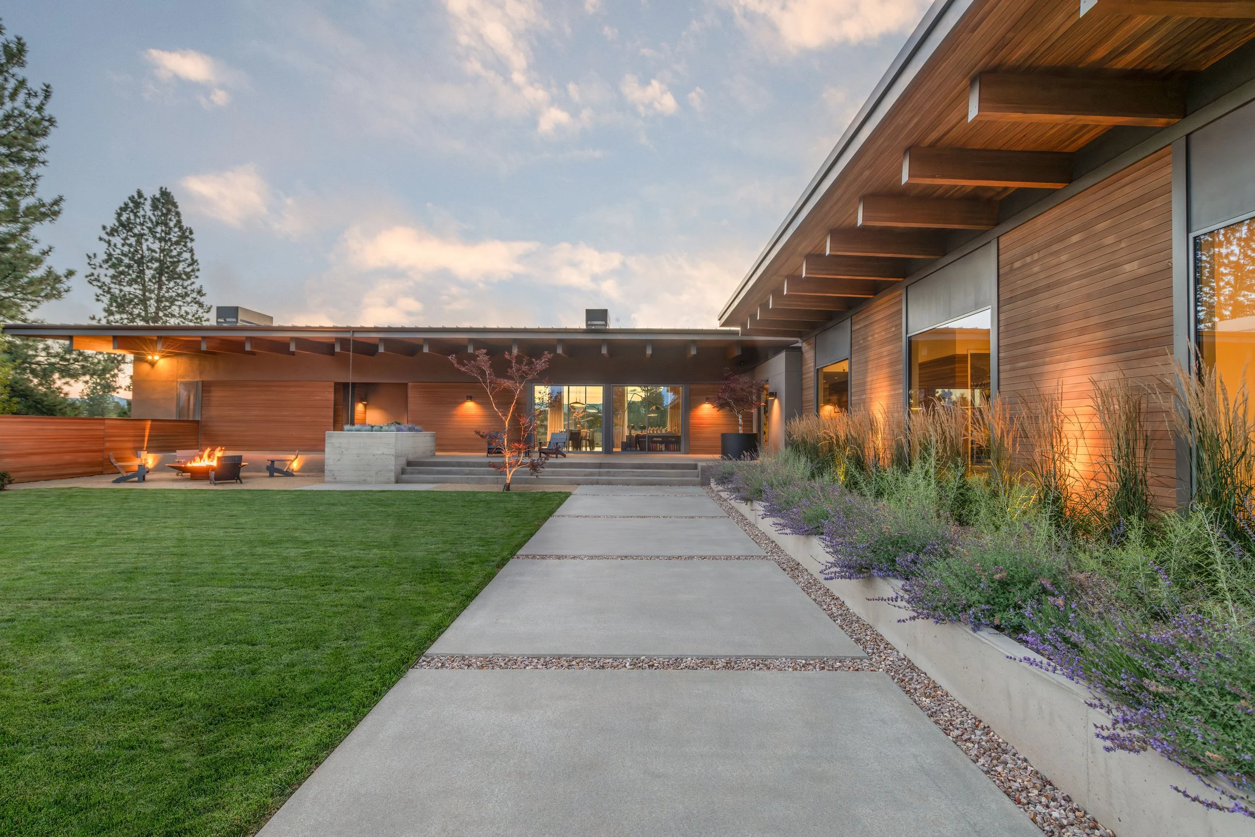 Modern custom home courtyard at dusk with cedar siding, exposed beam overhangs, concrete walkway, lavender landscaping, and fire pit in Bend, Oregon