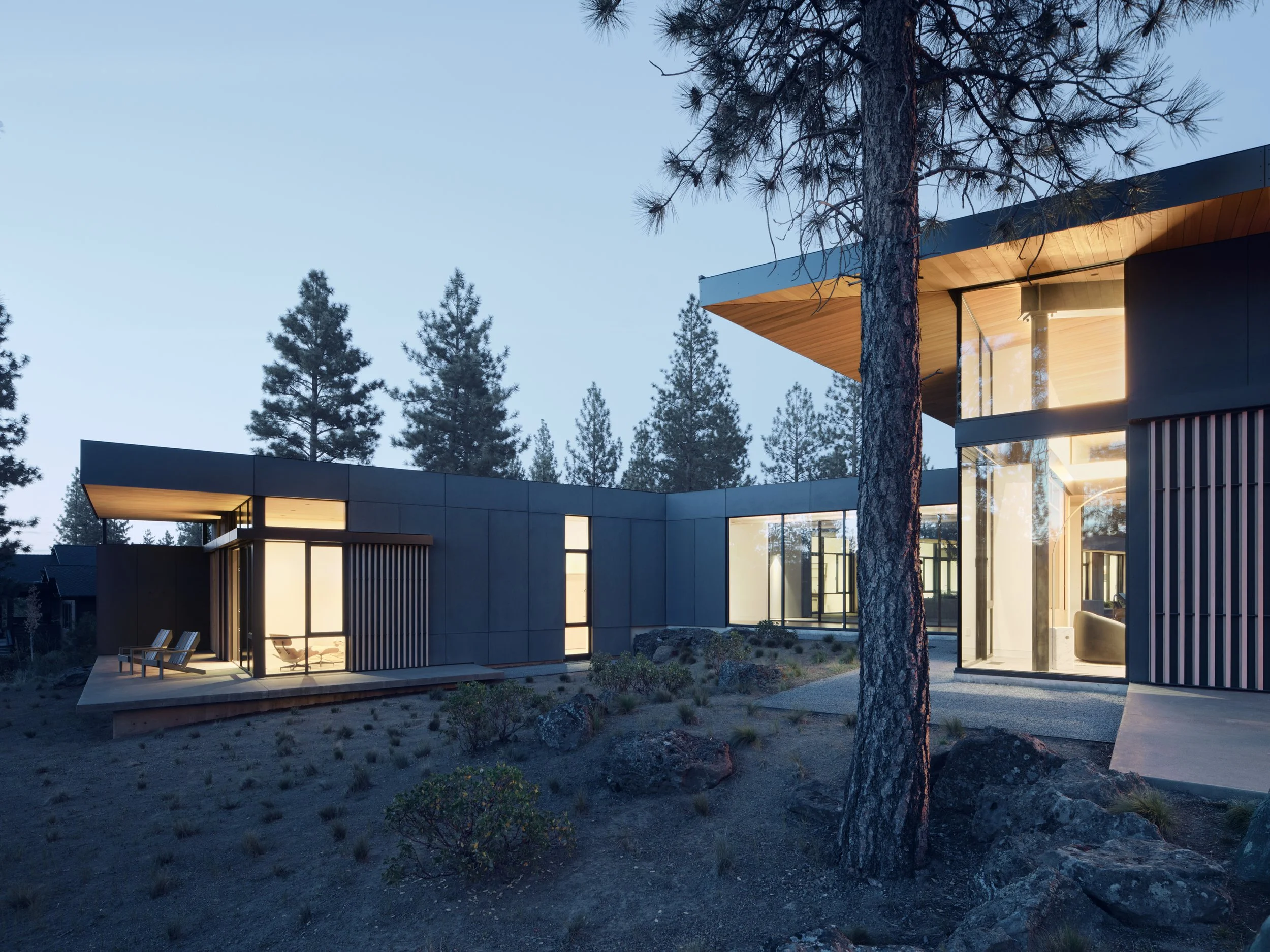 Modern house with large glass windows in a forest setting, lit from inside during dusk.