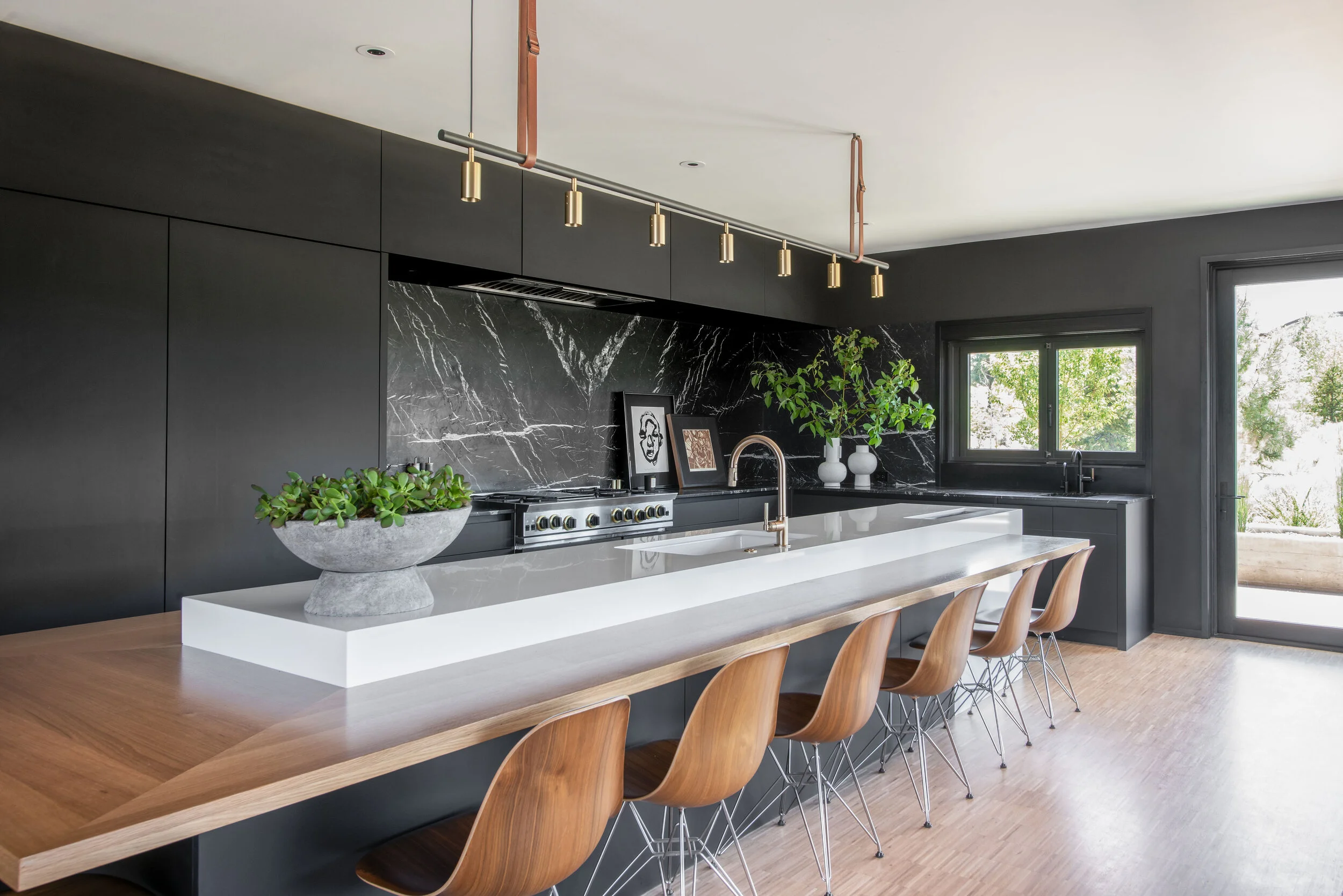 Modern kitchen with black cabinets, black marble backsplash, white countertop island, brown wooden chairs, plants, and a window with outdoor view.