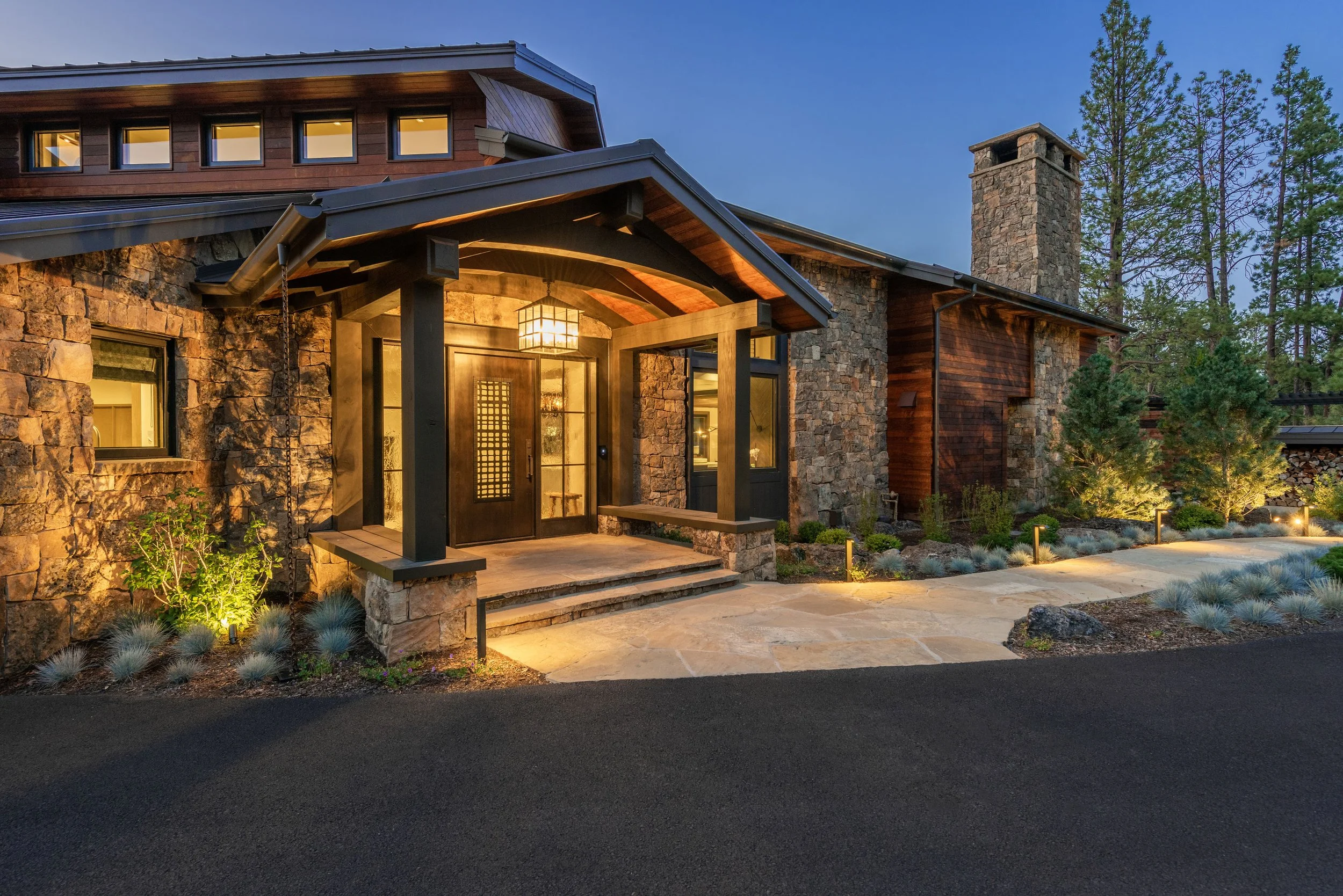 Front view of a modern house with stone and wood exterior, illuminated porch, and landscaped pathway, surrounded by tall pine trees at dusk.