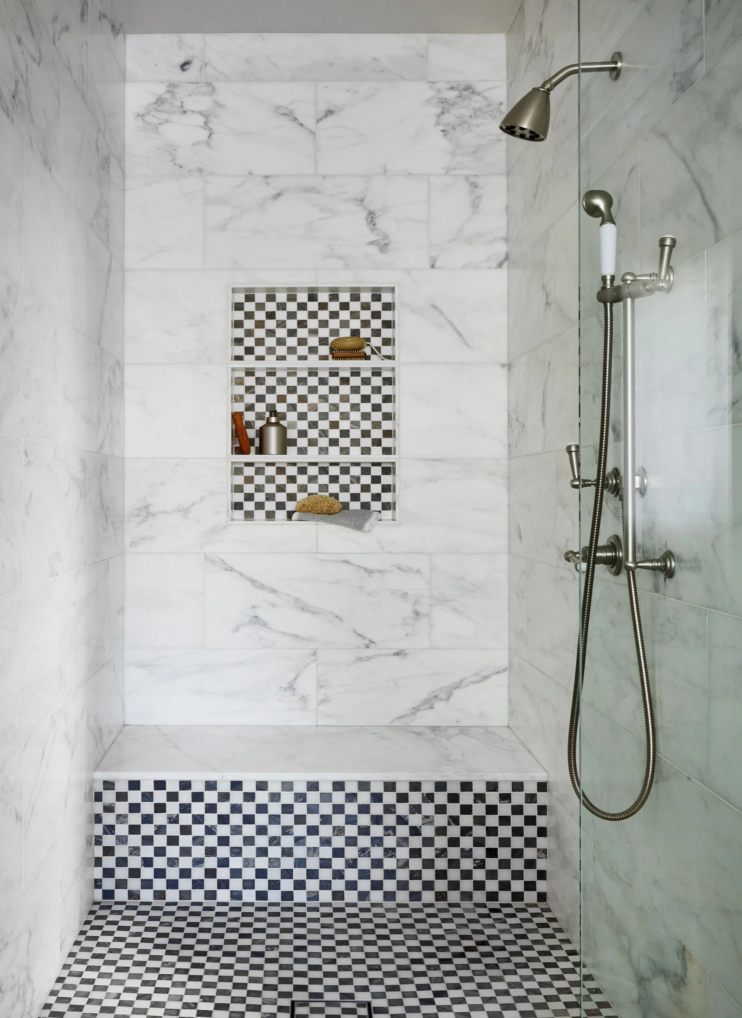 A marble-tiled walk-in shower with built-in shelves, a showerhead, a handheld shower, and decorative black-and-white checkered tiles on the shelves, floor, and bench.