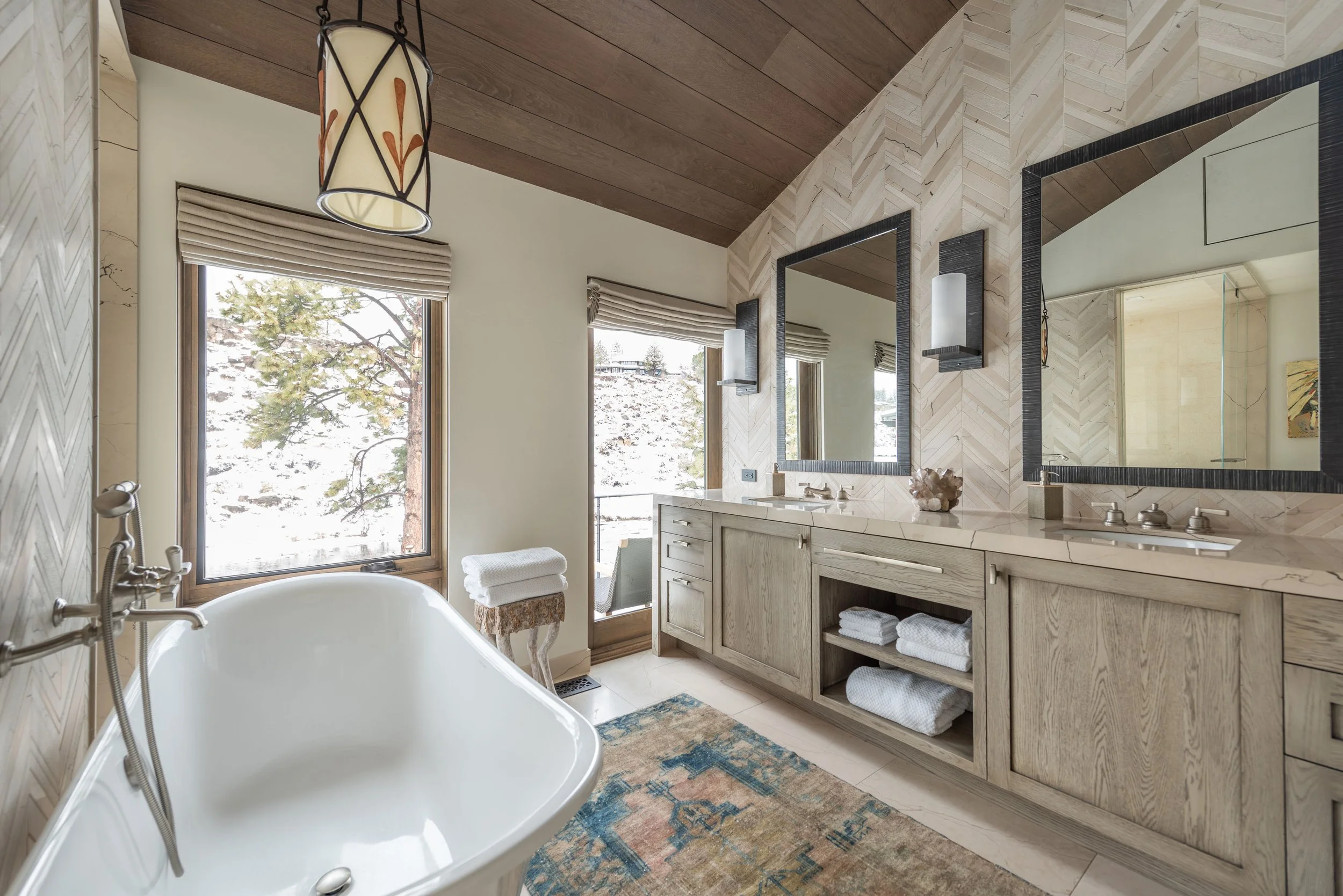 A spacious bathroom with a freestanding bathtub, two large mirrors above a wooden vanity, and large windows showing a snowy landscape outside.