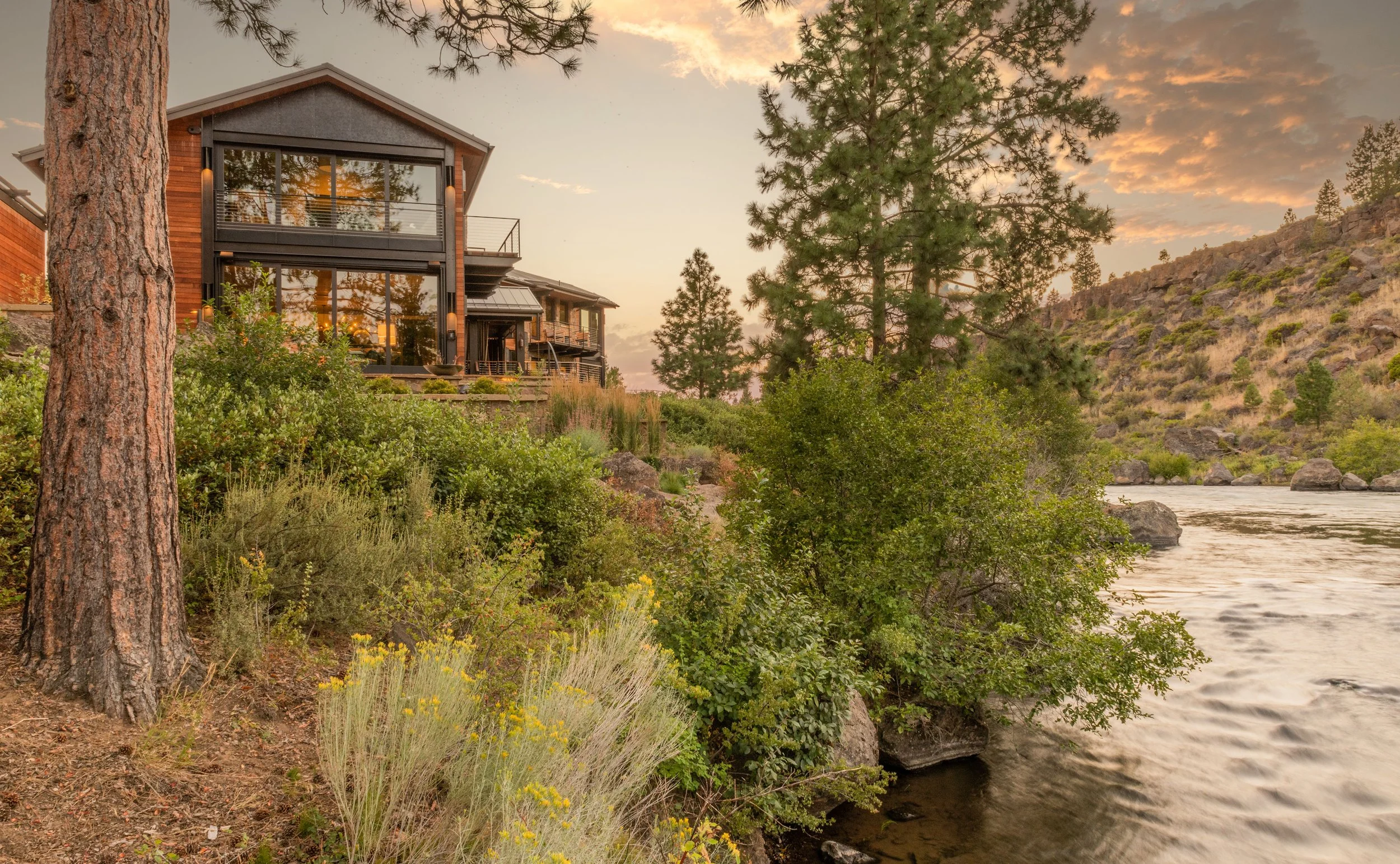 Modern house with large glass windows and outdoor deck overlooking a river, surrounded by trees and rocky hills at sunset.