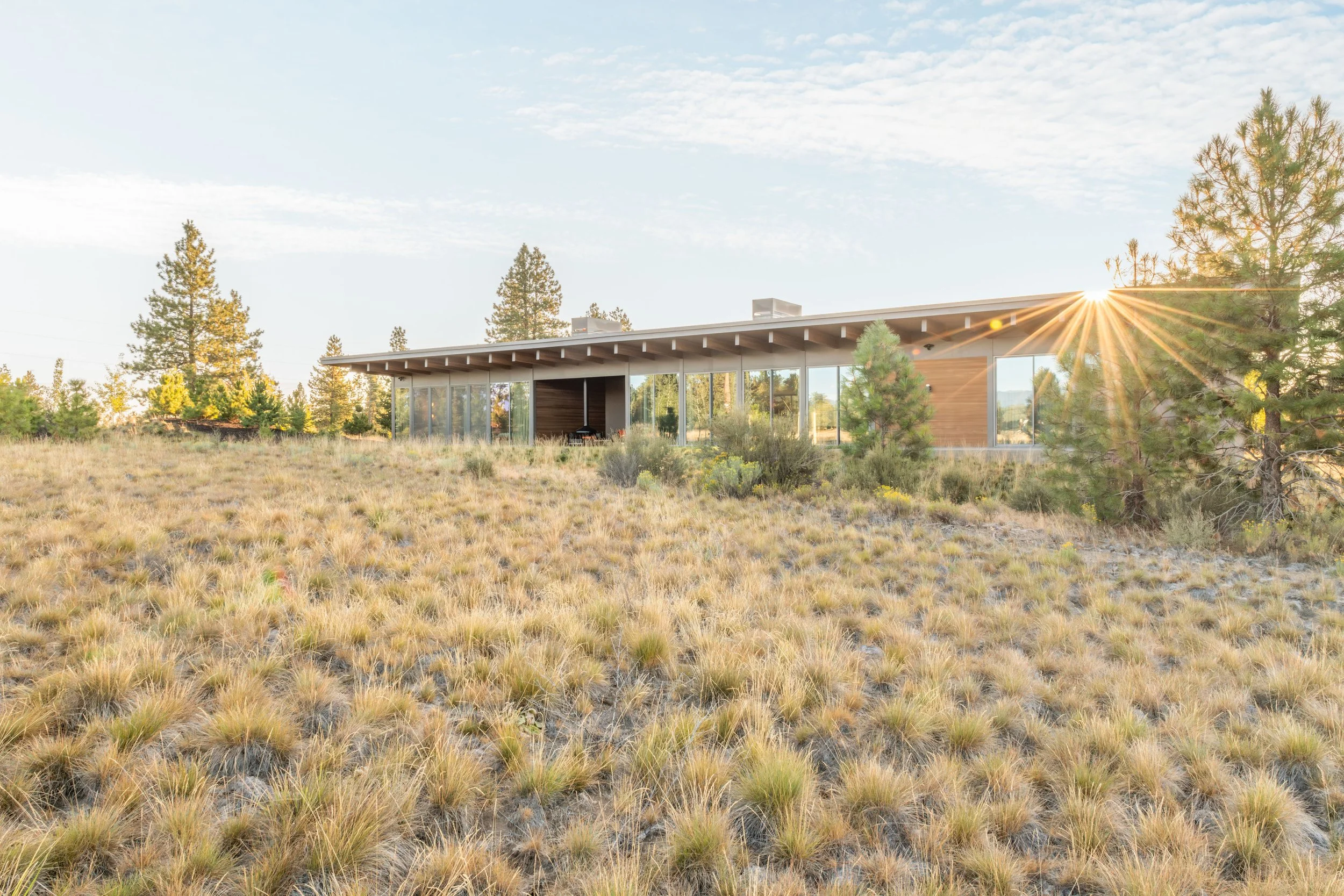 Wide exterior view of a single-story High Desert modern custom home in Bend, Oregon, featuring a flat roofline, extensive floor-to-ceiling windows, and native grassy landscaping bathed in golden sunset light.