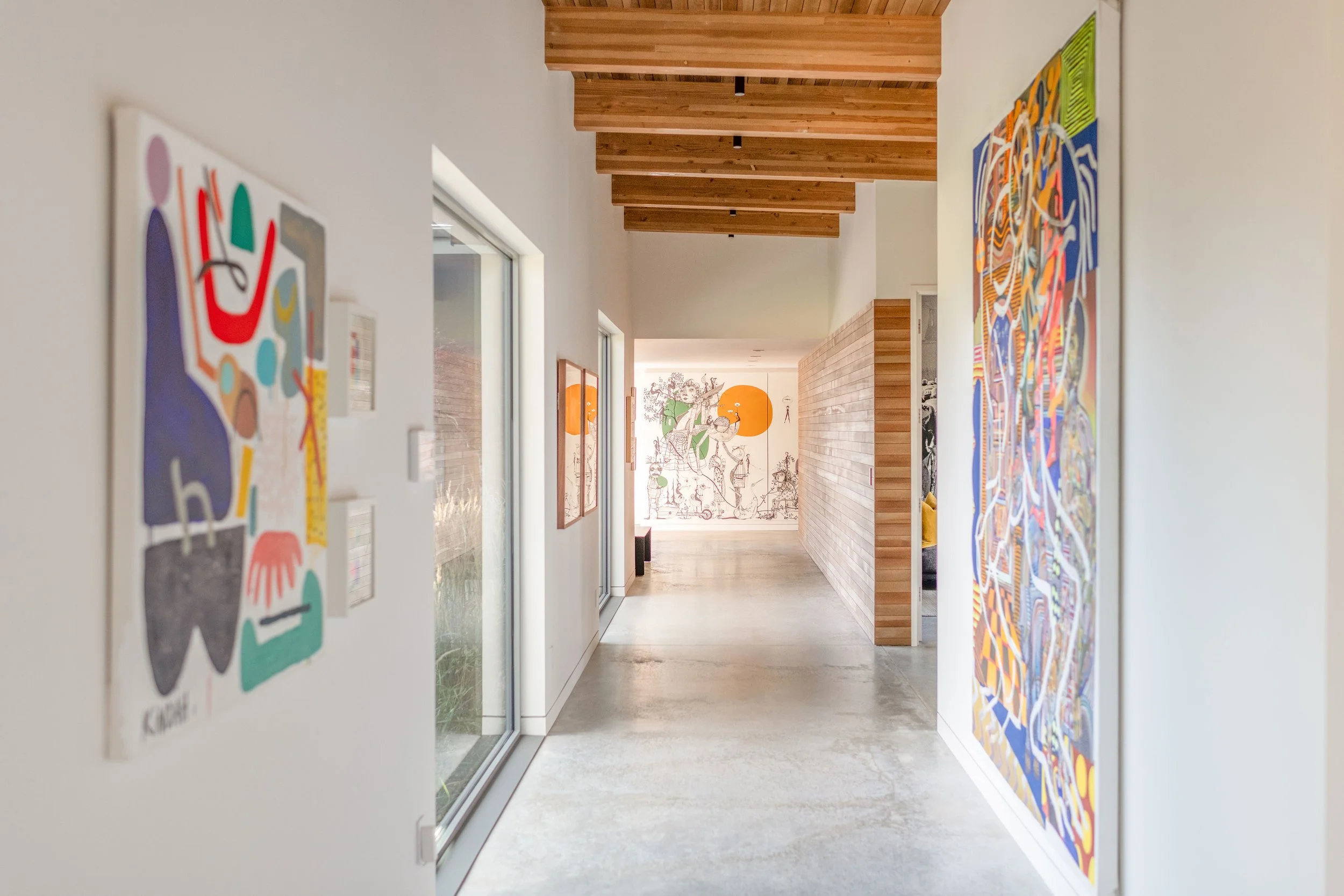 HoMA gallery hallway with polished concrete floors, wood beam ceiling, colorful contemporary artwork, and whimsical mural in Bend Oregon custom home