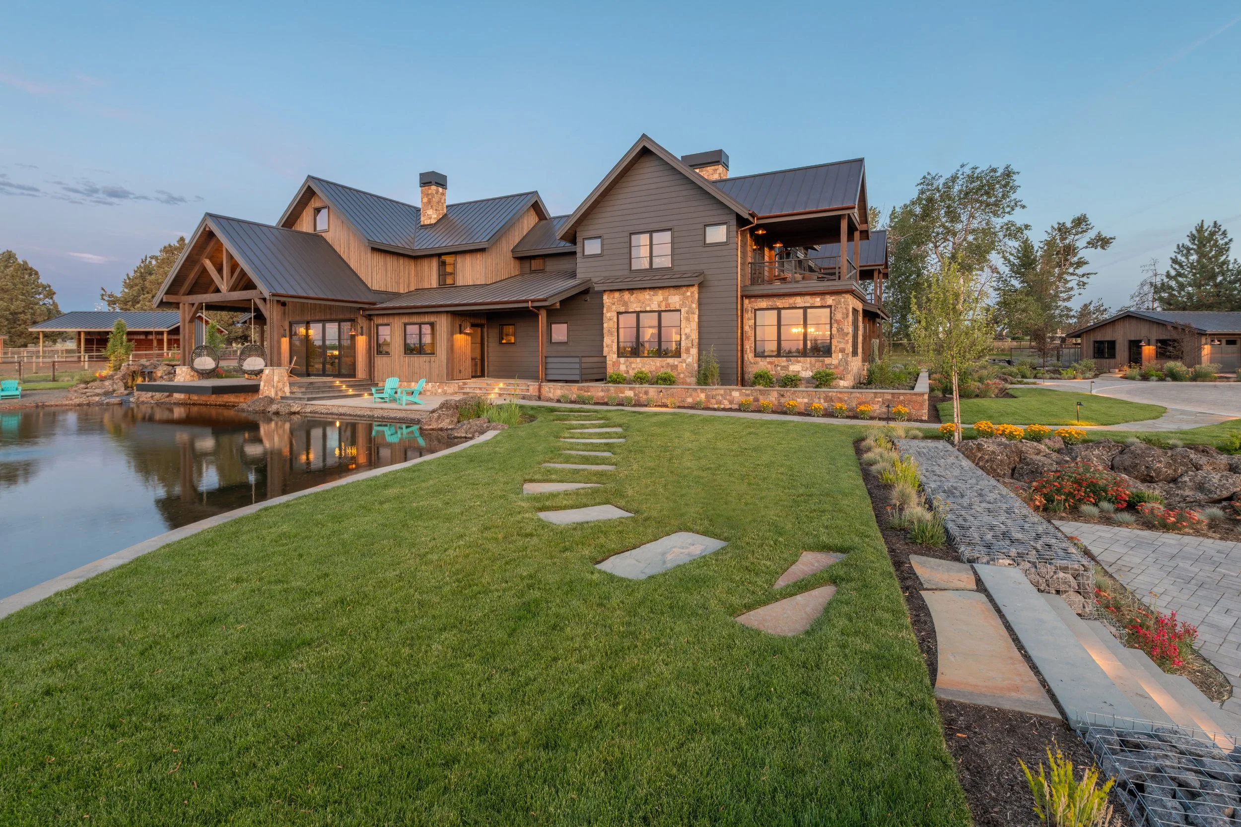 A large modern house with wood and stone exterior, surrounded by a manicured lawn, a pond, patio furniture, and landscaped gardens at sunset.