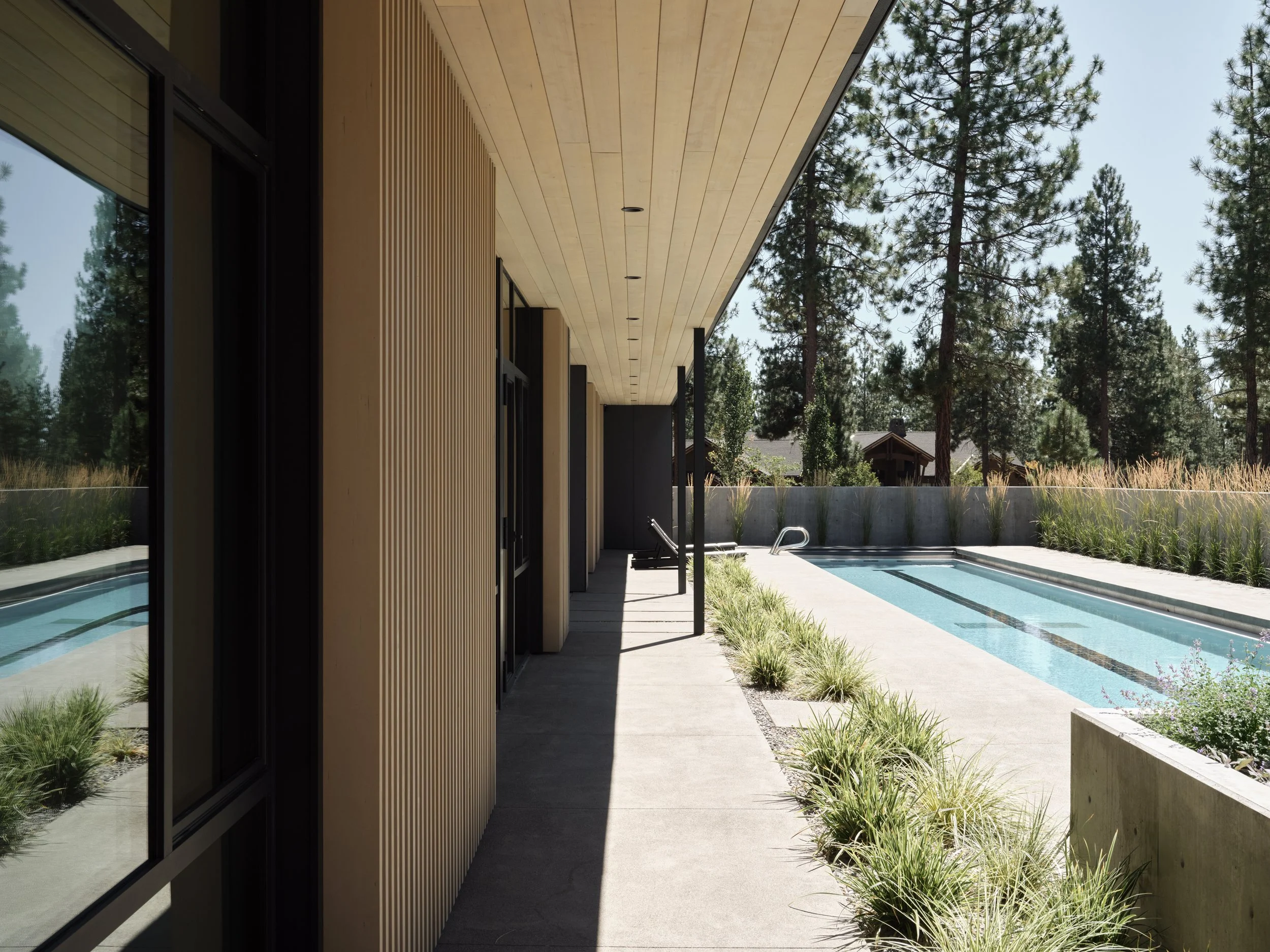 Side view of a modern house with a covered patio and a rectangular swimming pool, surrounded by trees and mountains in the background.
