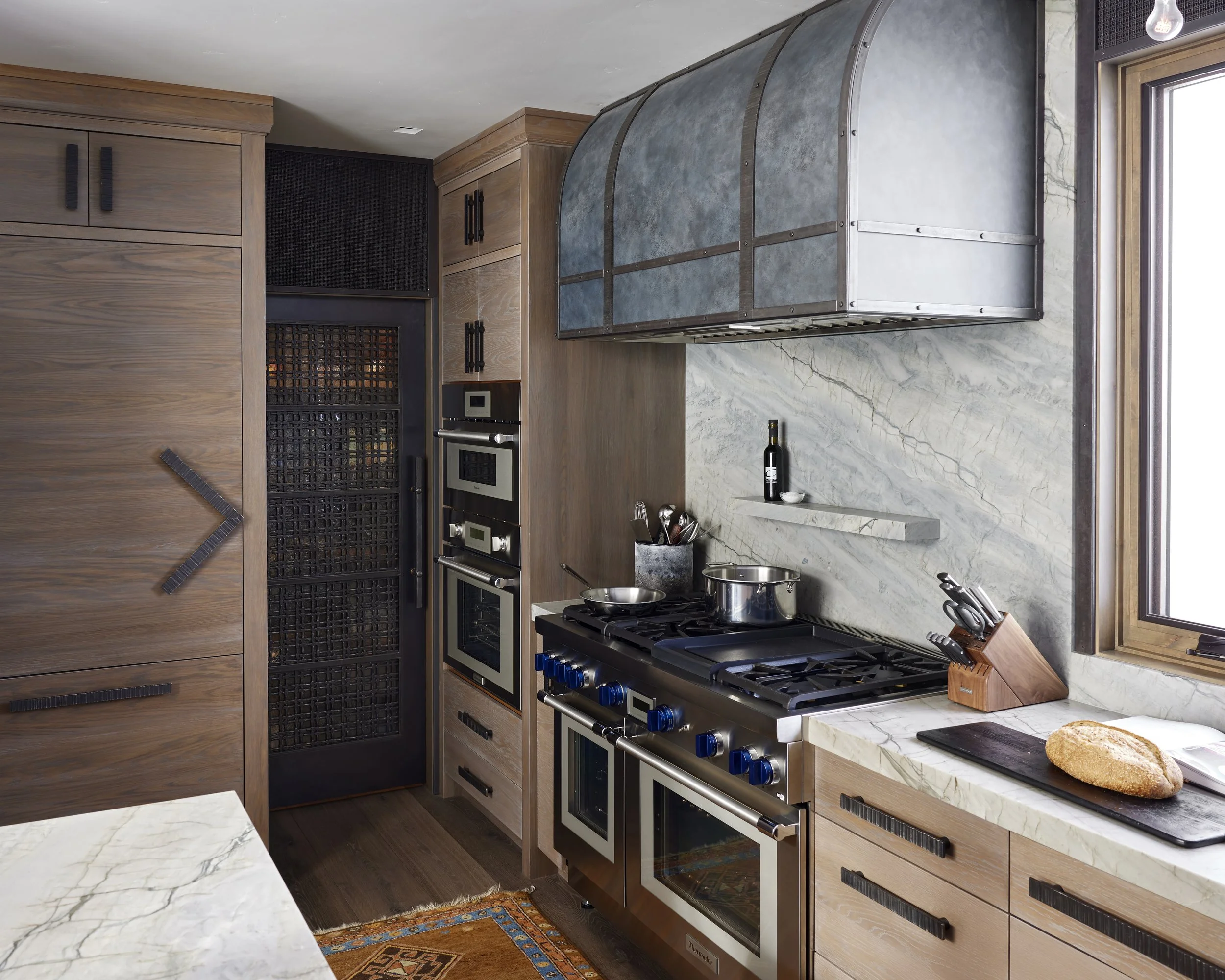 A modern kitchen with wood cabinetry, a marble backsplash, and stainless steel appliances including a stove, oven, and microwave. There is a loaf of bread on a black cutting board near a window.