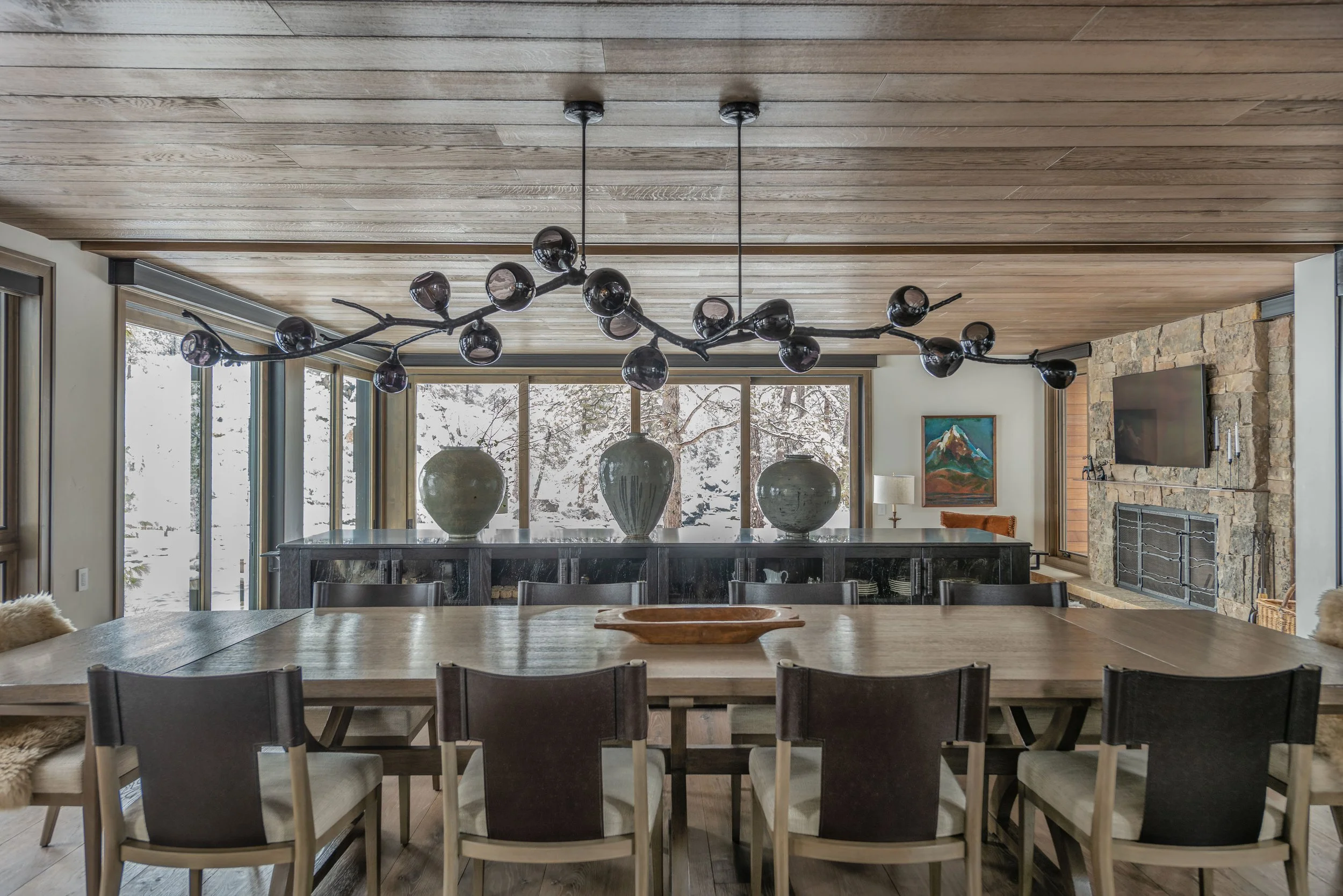 Spacious dining room with a large wooden table, multiple black chairs with beige cushions, and decorative large vases on a black sideboard against large windows showing snowy landscape outside. The ceiling is wood-paneled with a modern black chandeli