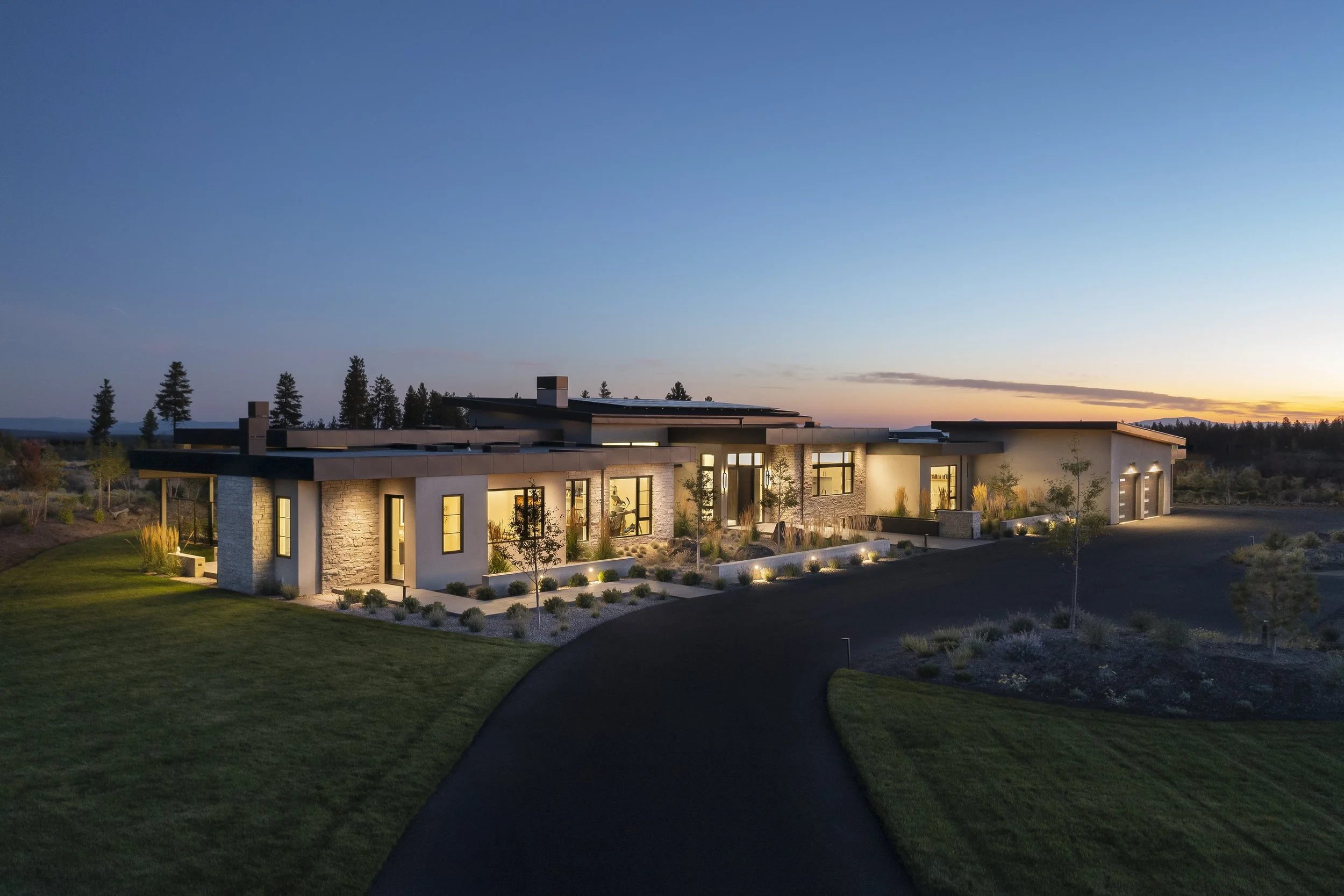 Panoramic twilight view of a luxury single-story custom home in Bend, Oregon, featuring modern flat-roof architecture, stone exterior cladding, and expansive windows glowing with warm interior light amidst high desert landscaping.