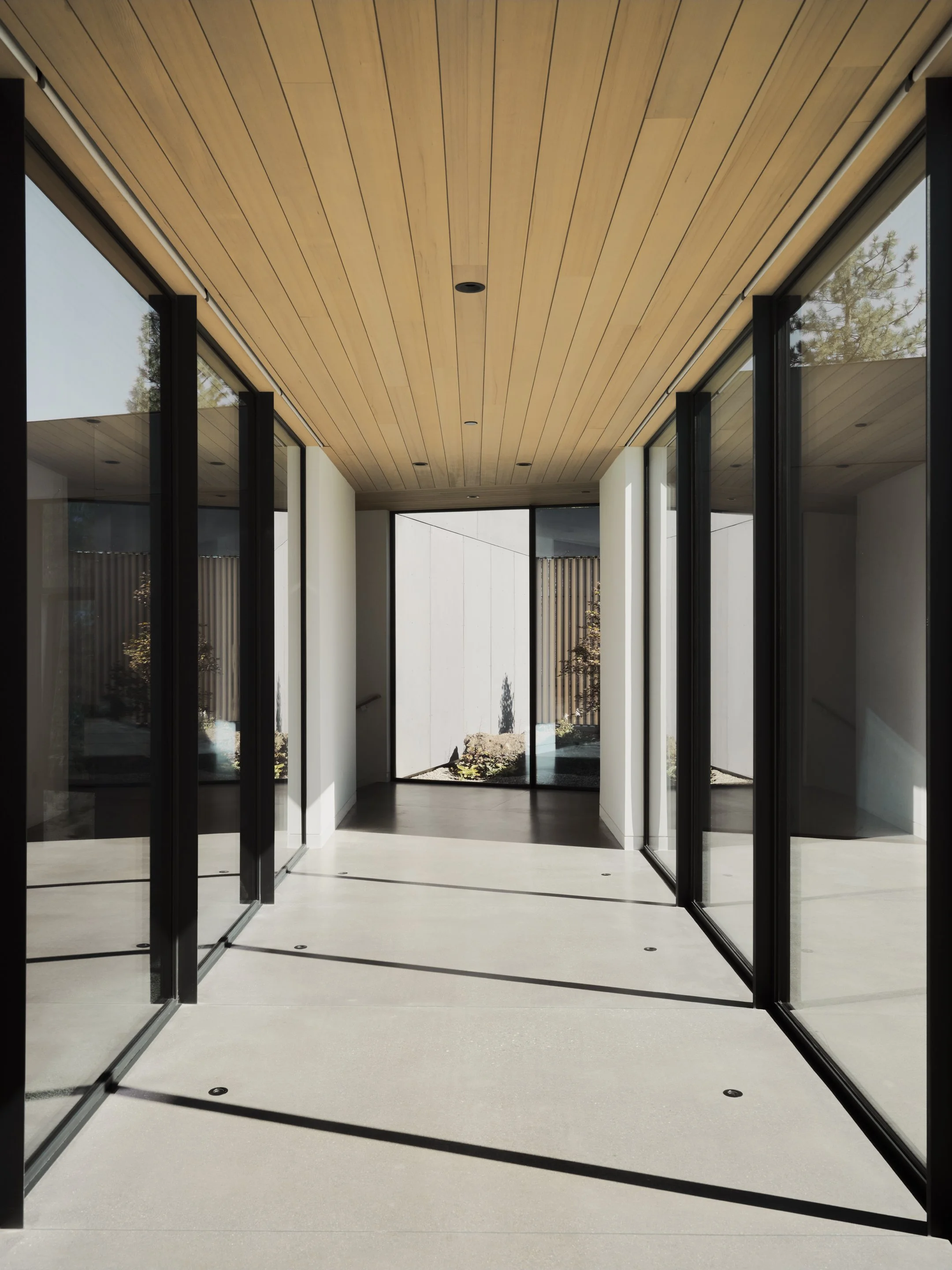 Modern hallway with glass walls, wooden ceiling, and a view of a garden outside.
