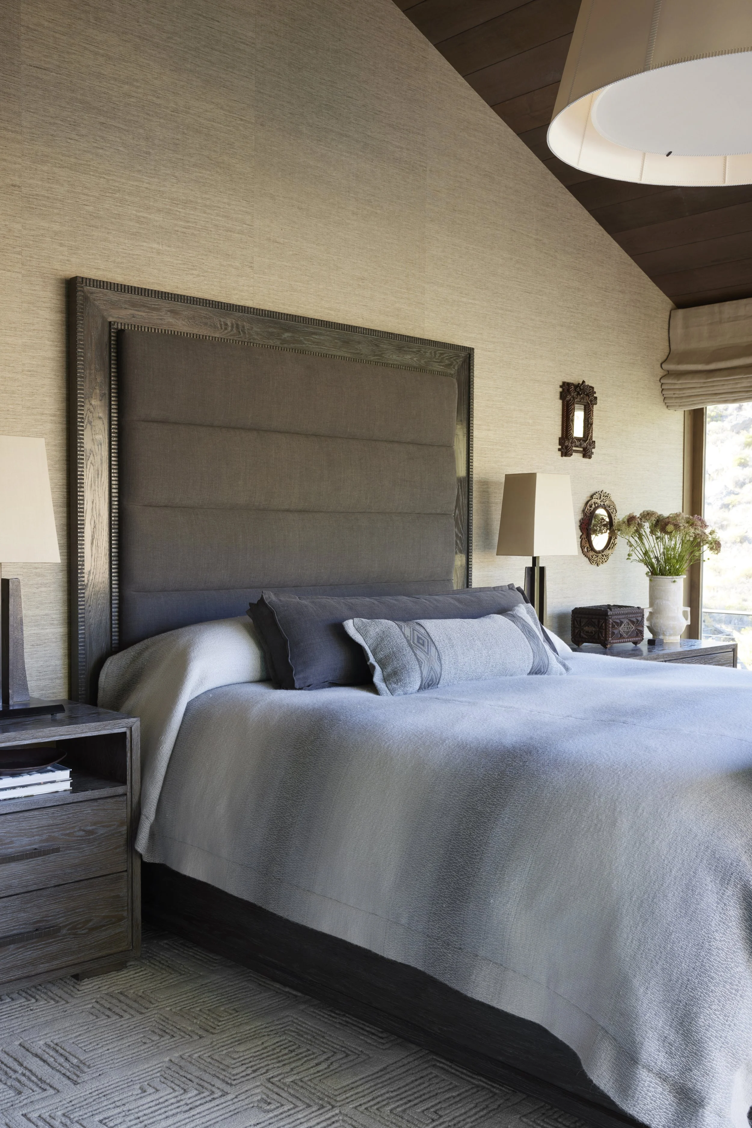 A neatly made bed with a tall headboard, pillows, and a throw blanket in a warmly lit bedroom with beige walls, wooden ceiling, side lamps, and a window with a view outside.