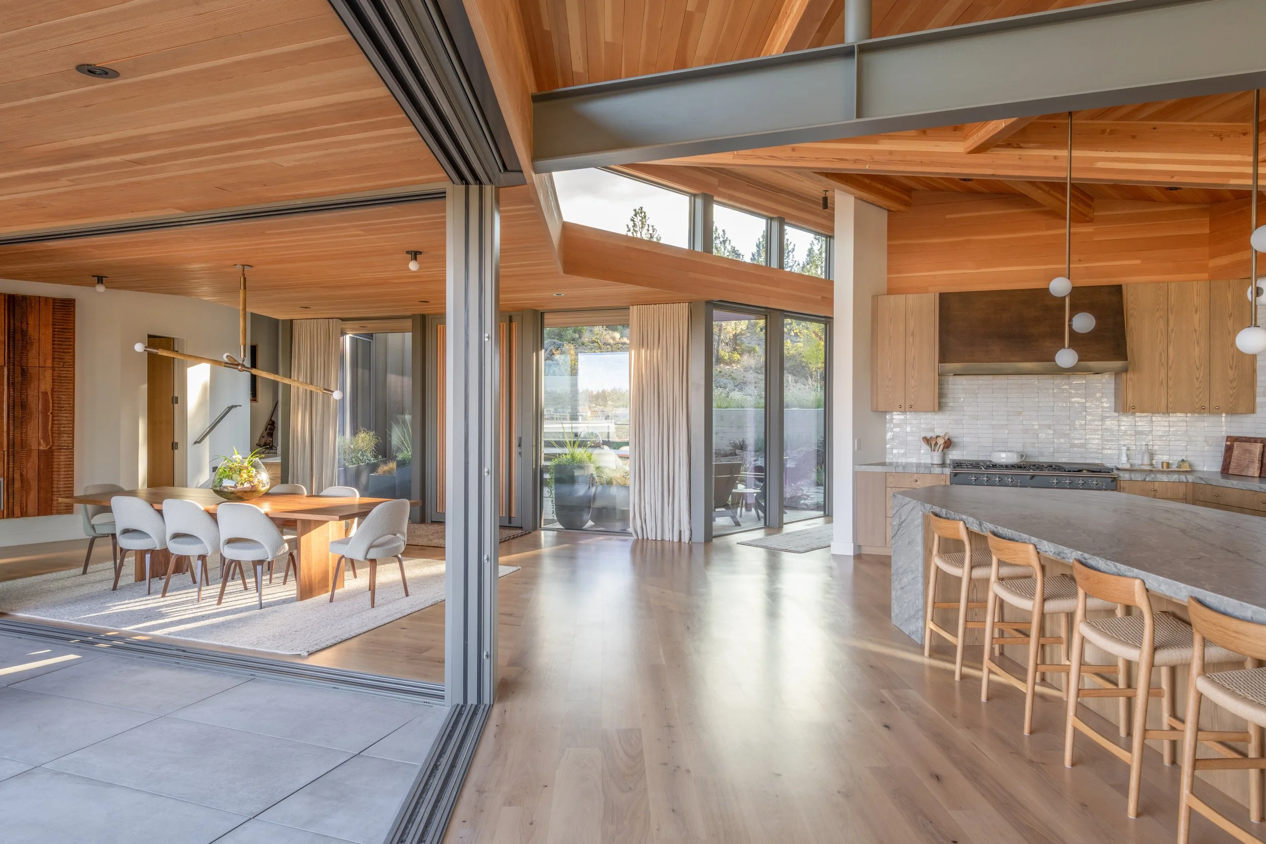 Open concept kitchen and dining room with wood beam ceiling, clerestory windows, stone waterfall island, and sliding glass doors to outdoor patio in Bend Oregon custom home