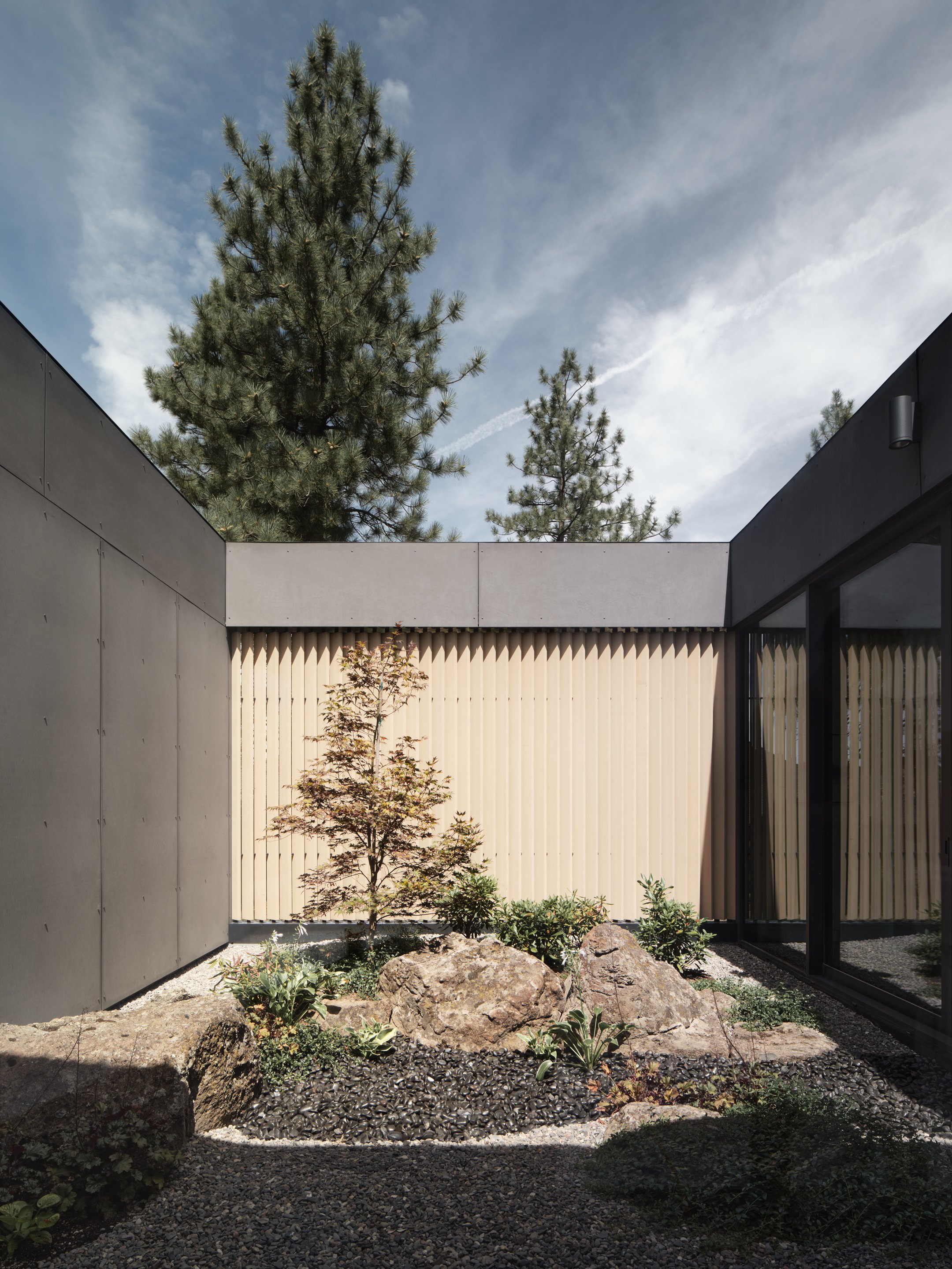 A modern outdoor courtyard with minimalist design features; a small garden bed with plants and rocks is enclosed by concrete and beige slatted wood walls, with tall pine trees visible in the background and a partly cloudy sky overhead.