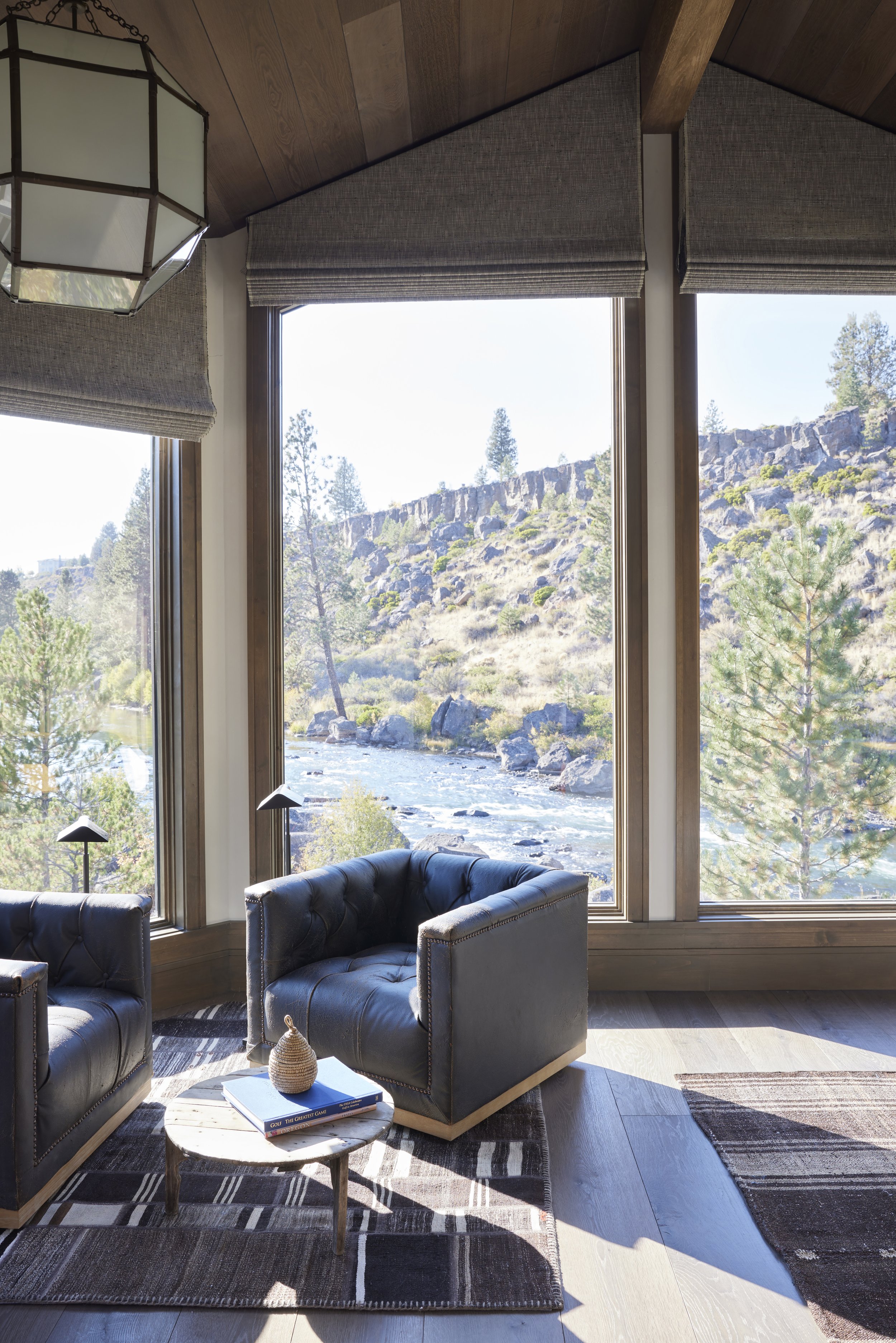 Living room with large windows overlooking a river and rocky hillside, featuring two black leather armchairs, a small round wooden table with books and a decorative object, a patterned rug, and a wooden ceiling with a geometric light fixture.