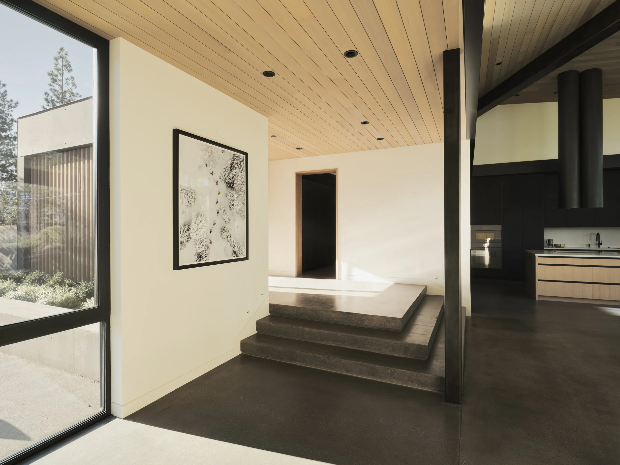 Modern interior with wooden ceiling, abstract artwork on cream wall, black staircase, black kitchen cabinets, and large window with outdoor greenery.