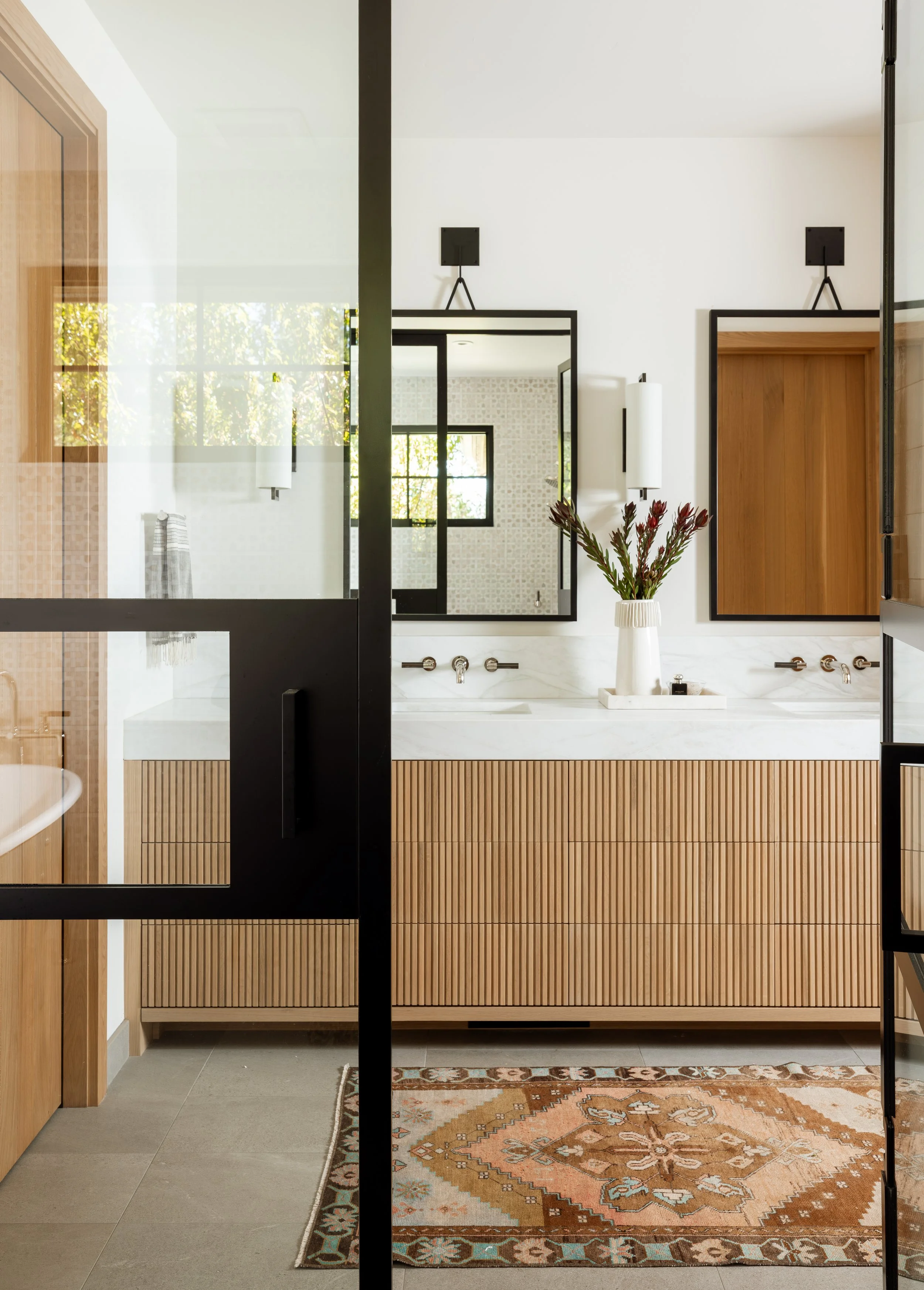 Master bathroom double vanity with a fluted wood front, seen through steel-framed glass.